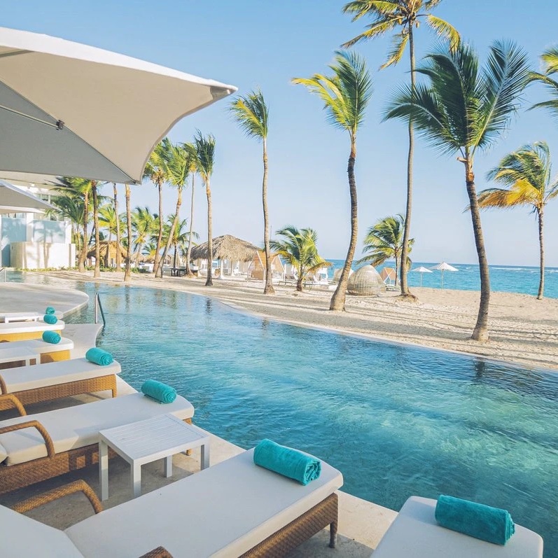 Luxury Iberostar Selection Bavaro perfect for a destination wedding, featuring an infinity pool, white sun loungers, turquoise towels, and a row of palm trees under a clear blue sky.