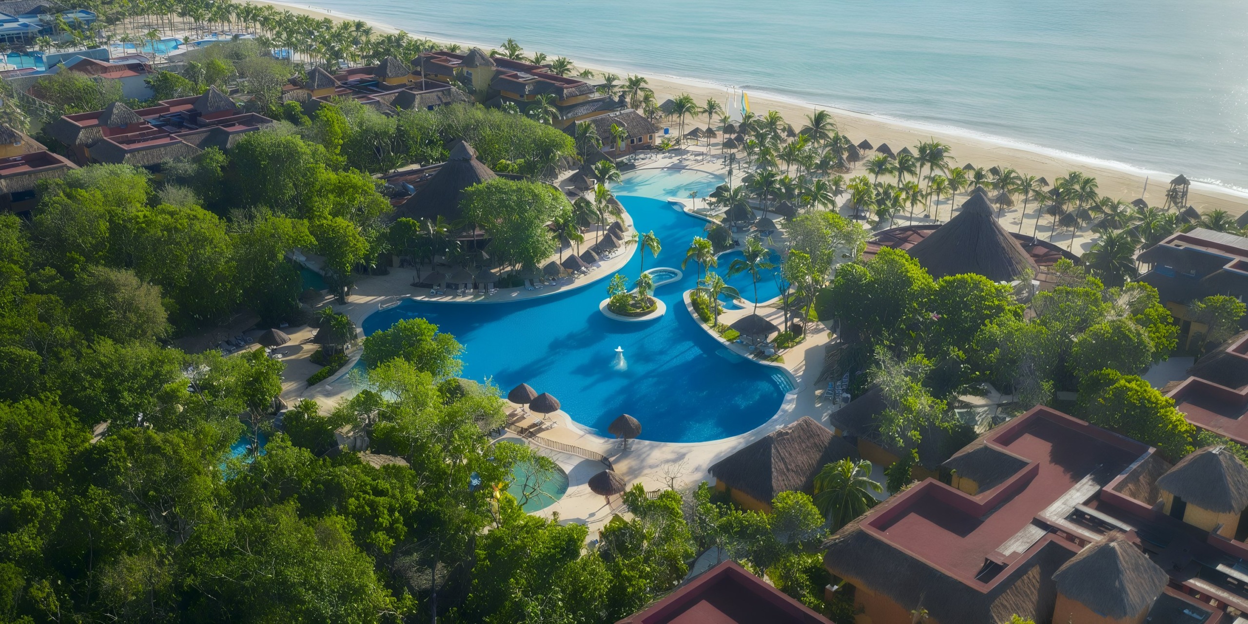 Aerial view of Iberostar Waves Quetzal, a resort with a large blue pool surrounded by tropical trees and thatched-roof buildings, adjacent to a sandy beach and ocean.