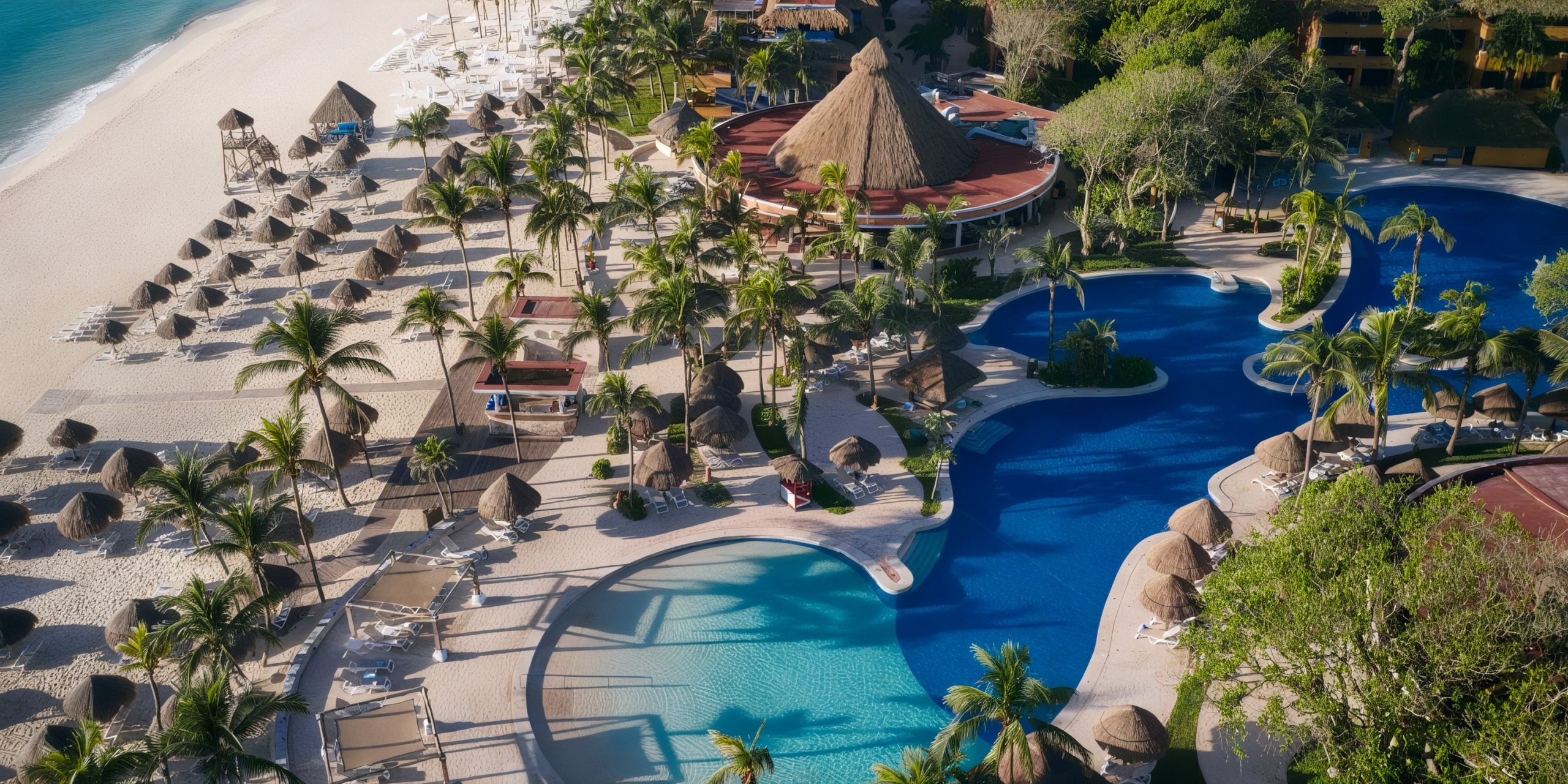 Aerial view of Iberostar Waves Quetzal, a tropical resort featuring multiple pools, palm trees, sun loungers, and beach huts along a white sandy beach.