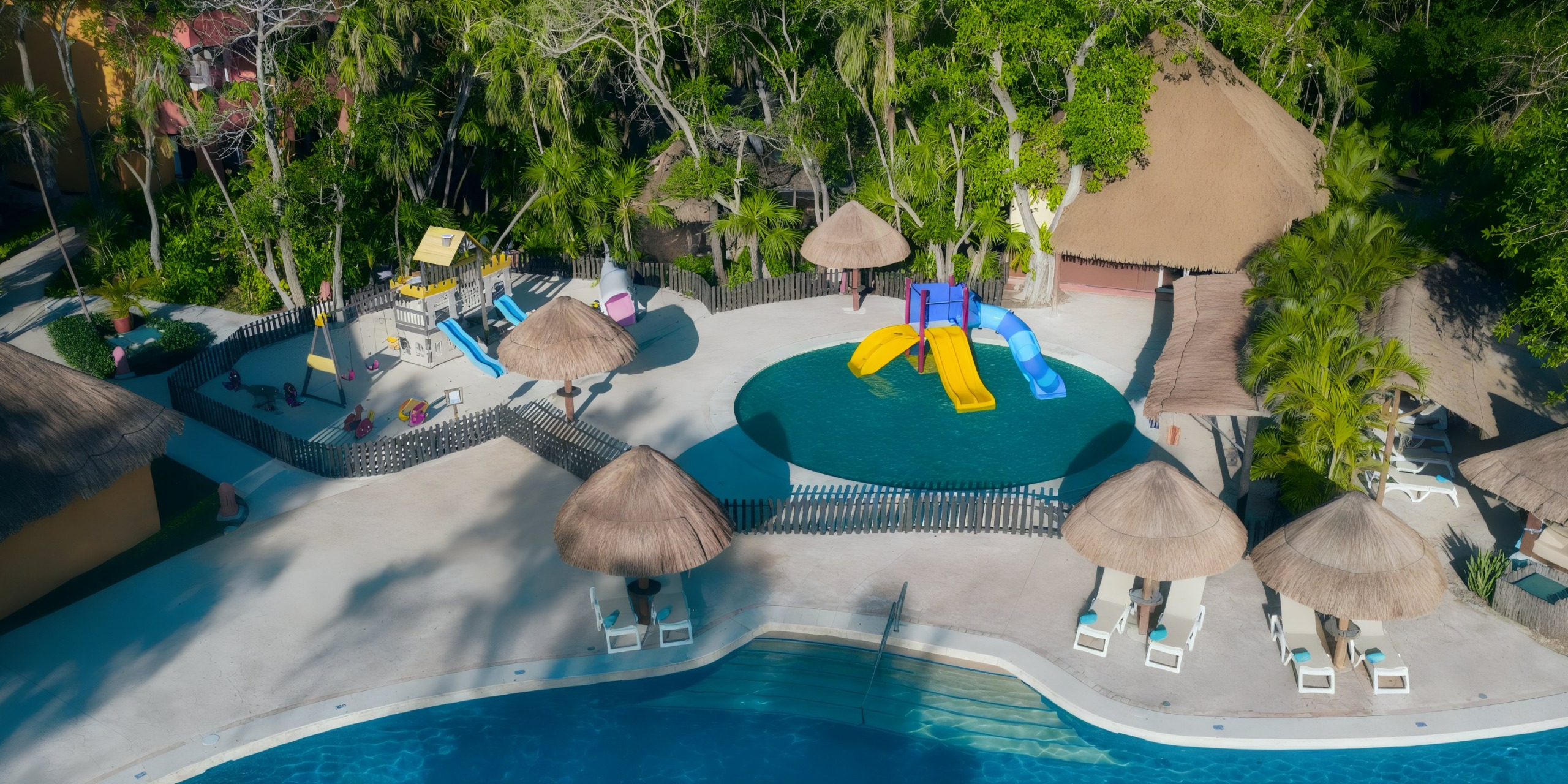 Aerial view of the Iberostar Waves Quetzal resort pool area with sun loungers, a children's splash pad featuring colorful slides, and thatched-roof structures surrounded by lush greenery.
