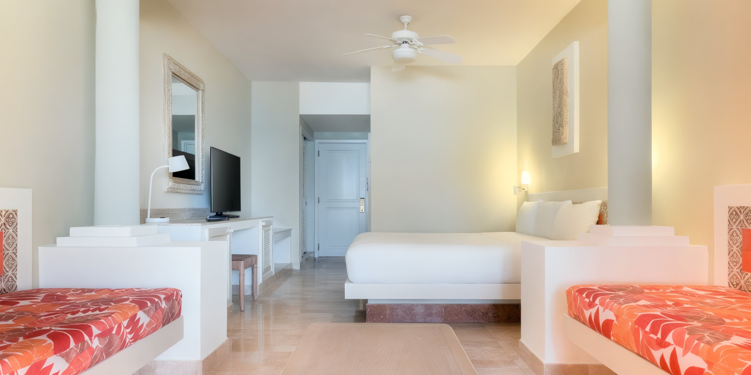 The Iberostar Waves Quetzal offers a hotel room featuring a king-size bed, red and white bench seating, a desk with a TV, a ceiling fan, and a white door. The decor is minimalistic in beige and white tones.