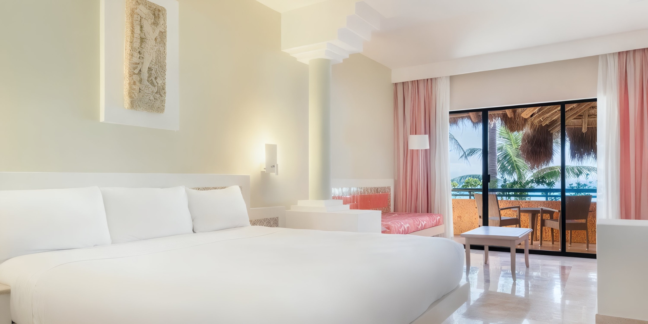 Experience the Iberostar Waves Quetzal, featuring a vibrant hotel room with a spacious bed, pink accent chairs, a table, and a balcony offering views of palm trees and the ocean.