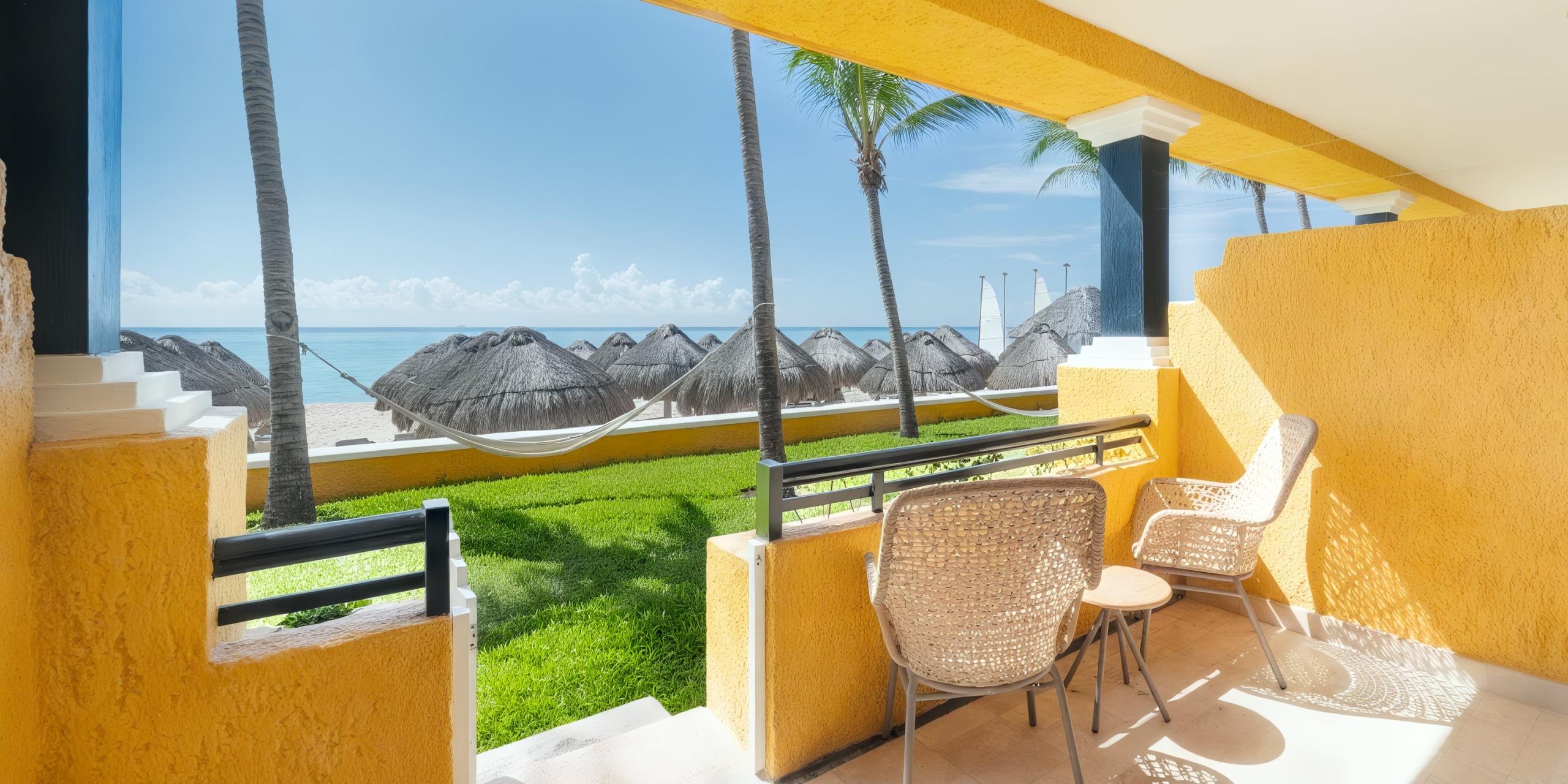 The Iberostar Waves Quetzal features a patio with two chairs and a small table, offering views of a beach adorned with thatched umbrellas and hammocks, all set against the backdrop of palm trees and the ocean.