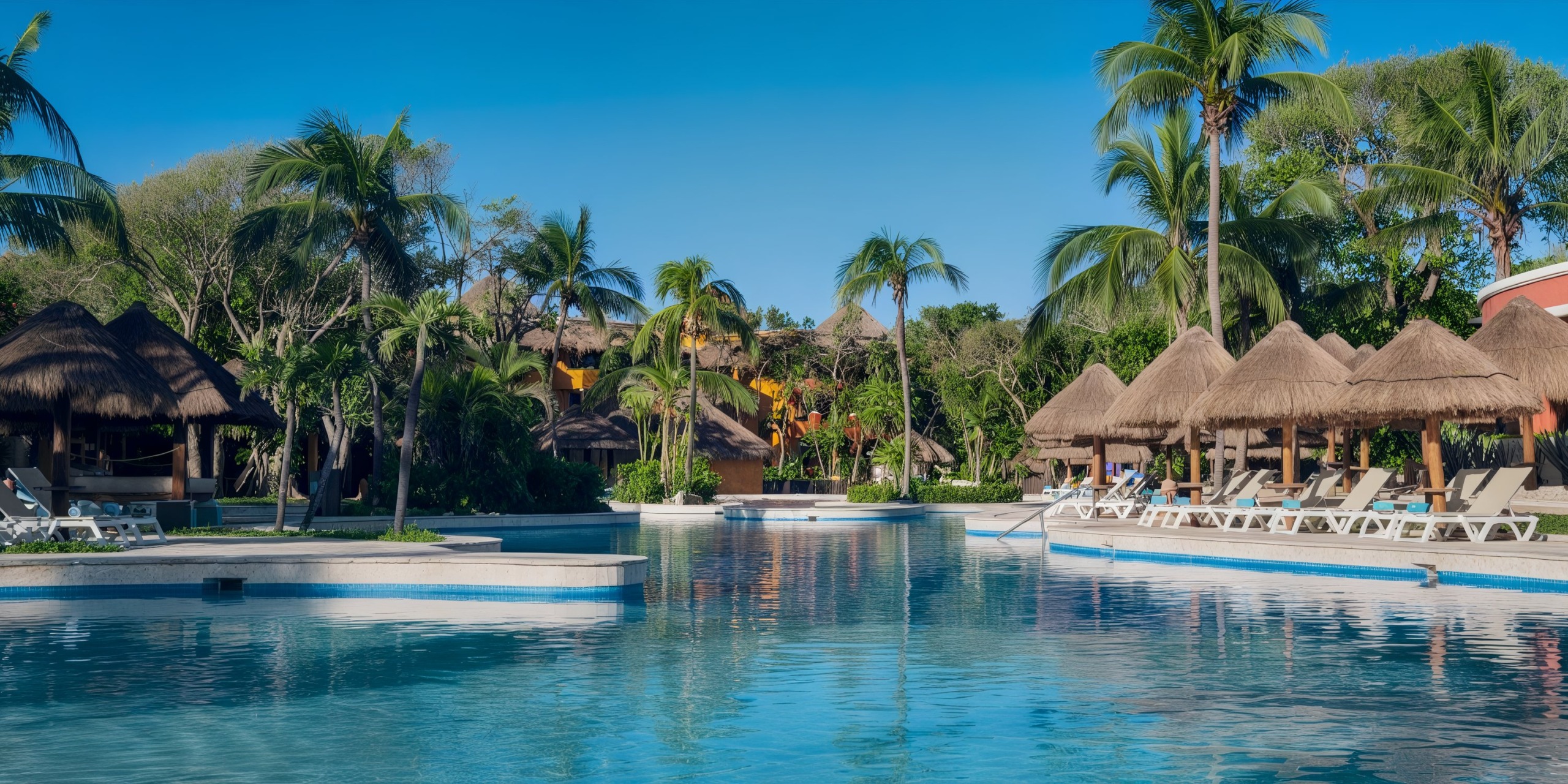 The Iberostar Waves Quetzal resort features a stunning pool area equipped with sun loungers and charming thatched umbrellas, all beautifully surrounded by lush palm trees under a clear blue sky.