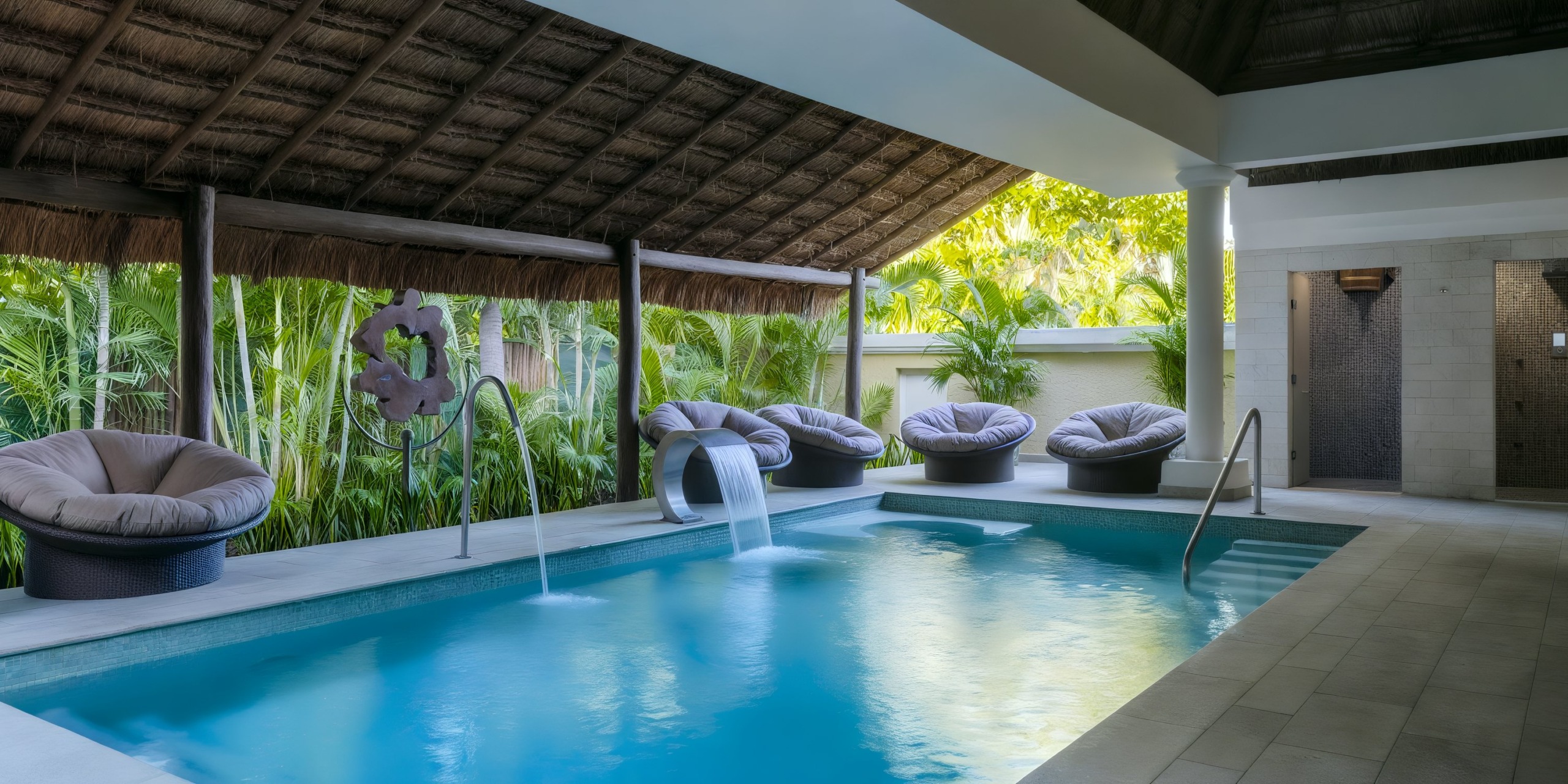 The Iberostar Waves Quetzal offers an indoor pool complete with cascading water features, encircled by cozy round lounge chairs under a thatched roof, all set against a backdrop of lush greenery.