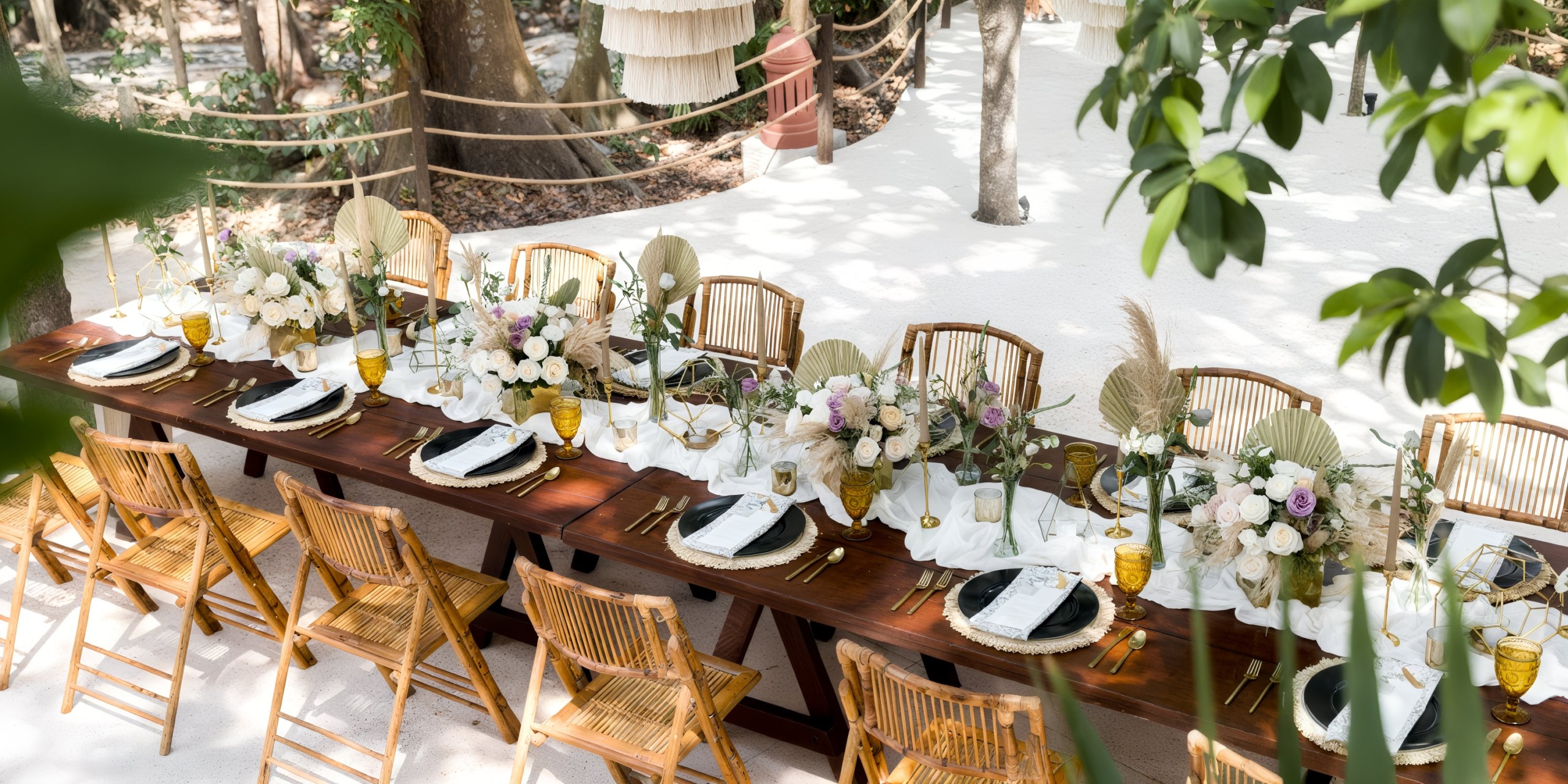 Iberostar Waves Quetzal outdoor dining set with wicker chairs, elegantly arranged with floral centerpieces, plates, and utensils on a white cloth backdrop. Enveloped by lush greenery and trees.