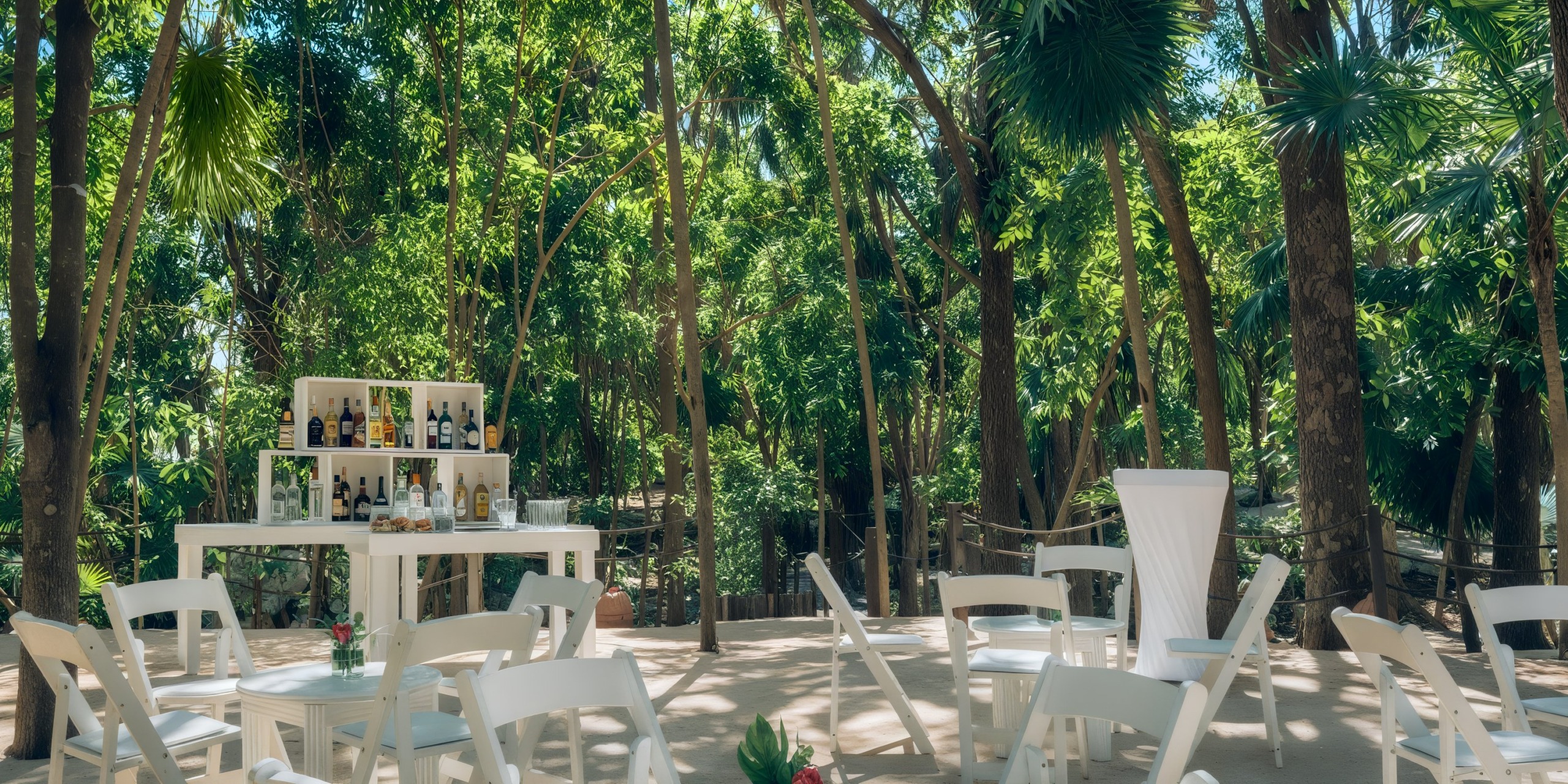 The Iberostar Waves Quetzal features an outdoor setup with white chairs and tables nestled in a forested area, with a bar stocked with various bottles in the background.