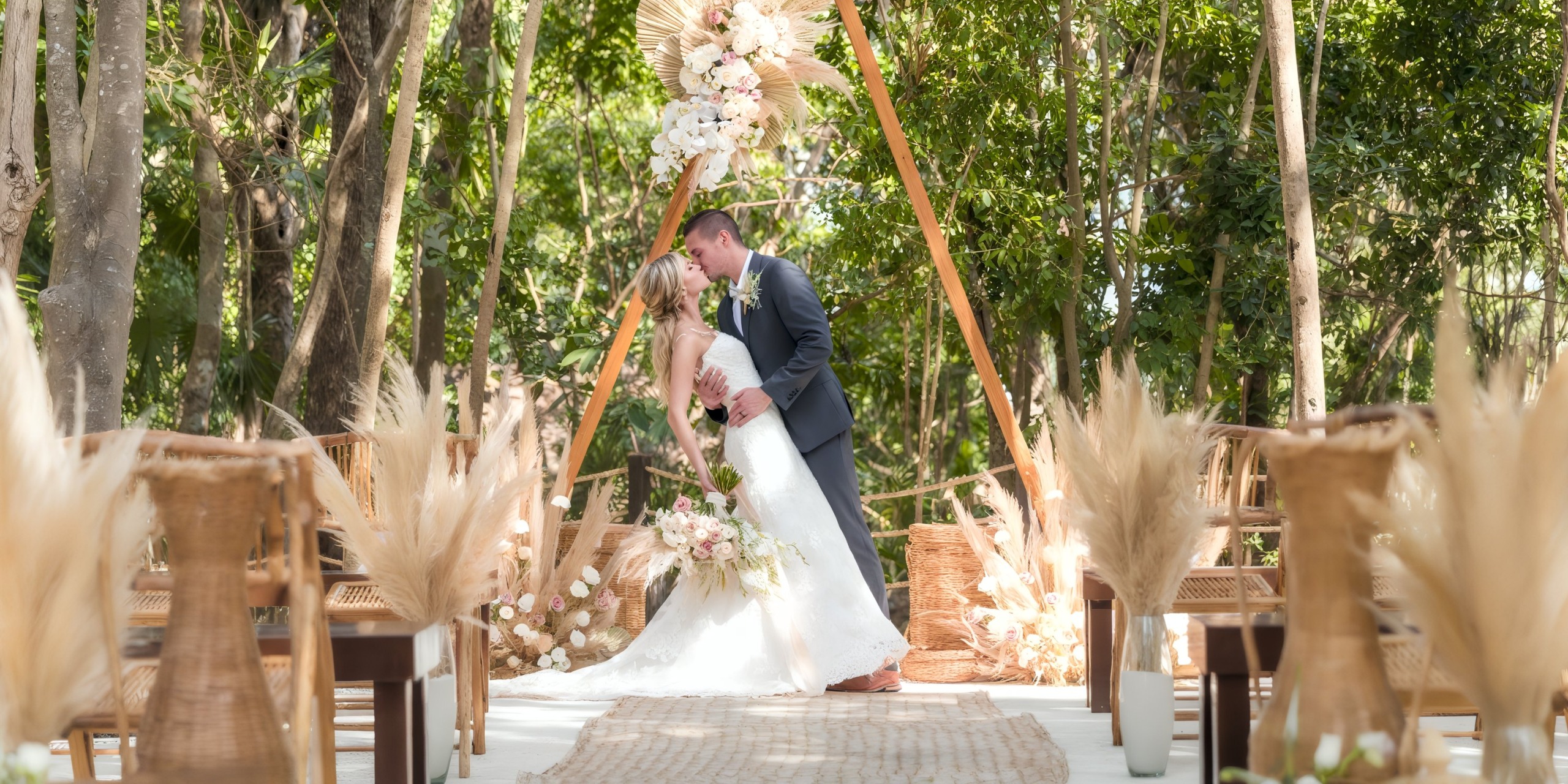 A couple in Iberostar Waves Quetzal wedding attire shares a kiss under a floral arch in a forest setting, with the aisle lined with wooden chairs adorned with pampas grass.
