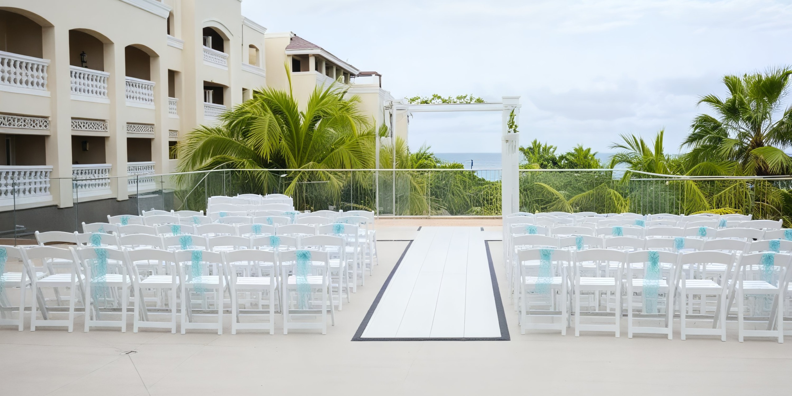 The Iberostar Waves Rose Hall Beach is beautifully arranged for a destination wedding, featuring an elegant setup with white chairs and an archway surrounded by lush greenery.