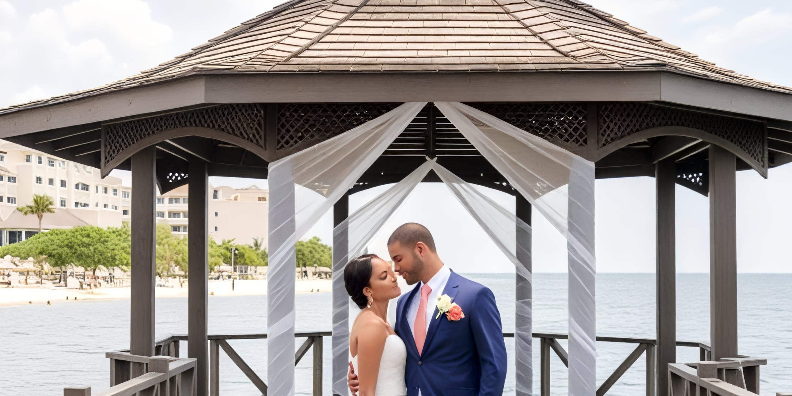 A couple at the Iberostar Waves Rose Hall Beach embraces by the sea under a gazebo, setting a picturesque scene for a destination wedding.