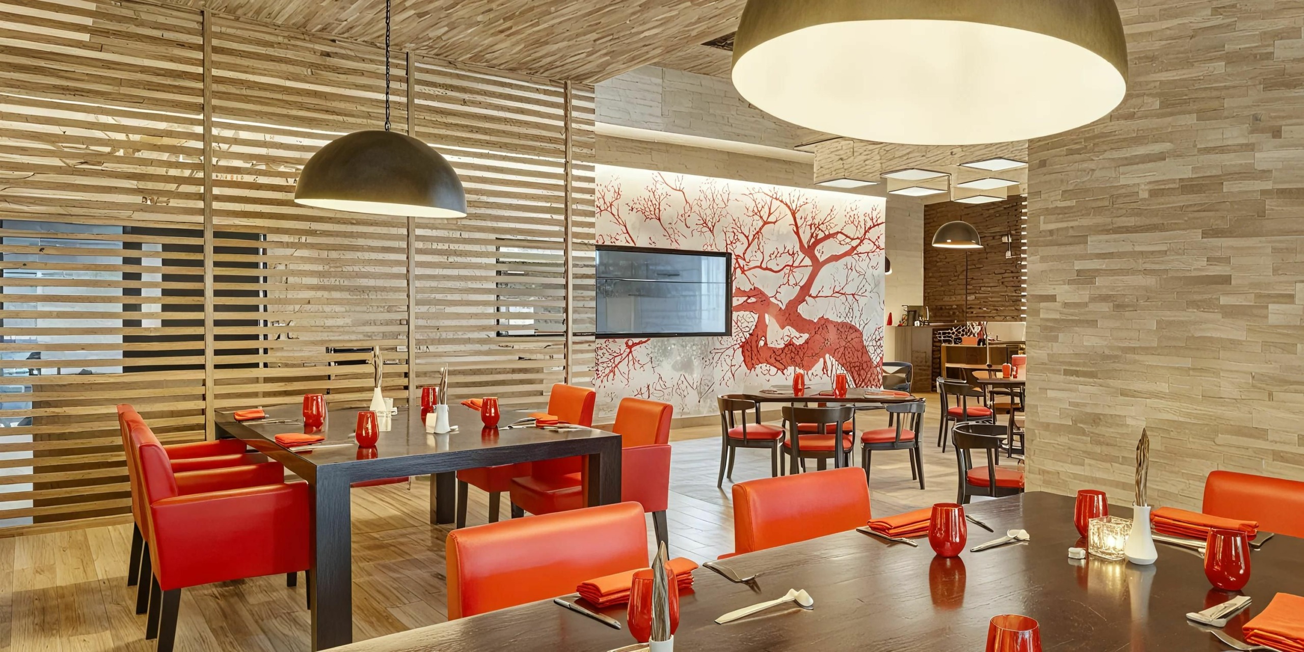 The Royalton Punta Cana features a modern restaurant interior adorned with wooden decor, vibrant red chairs, and a striking mural of a red tree. The tables are meticulously set with red napkins, pristine utensils, and elegant glasses. Large overhead lights bathe the space in warm illumination.
