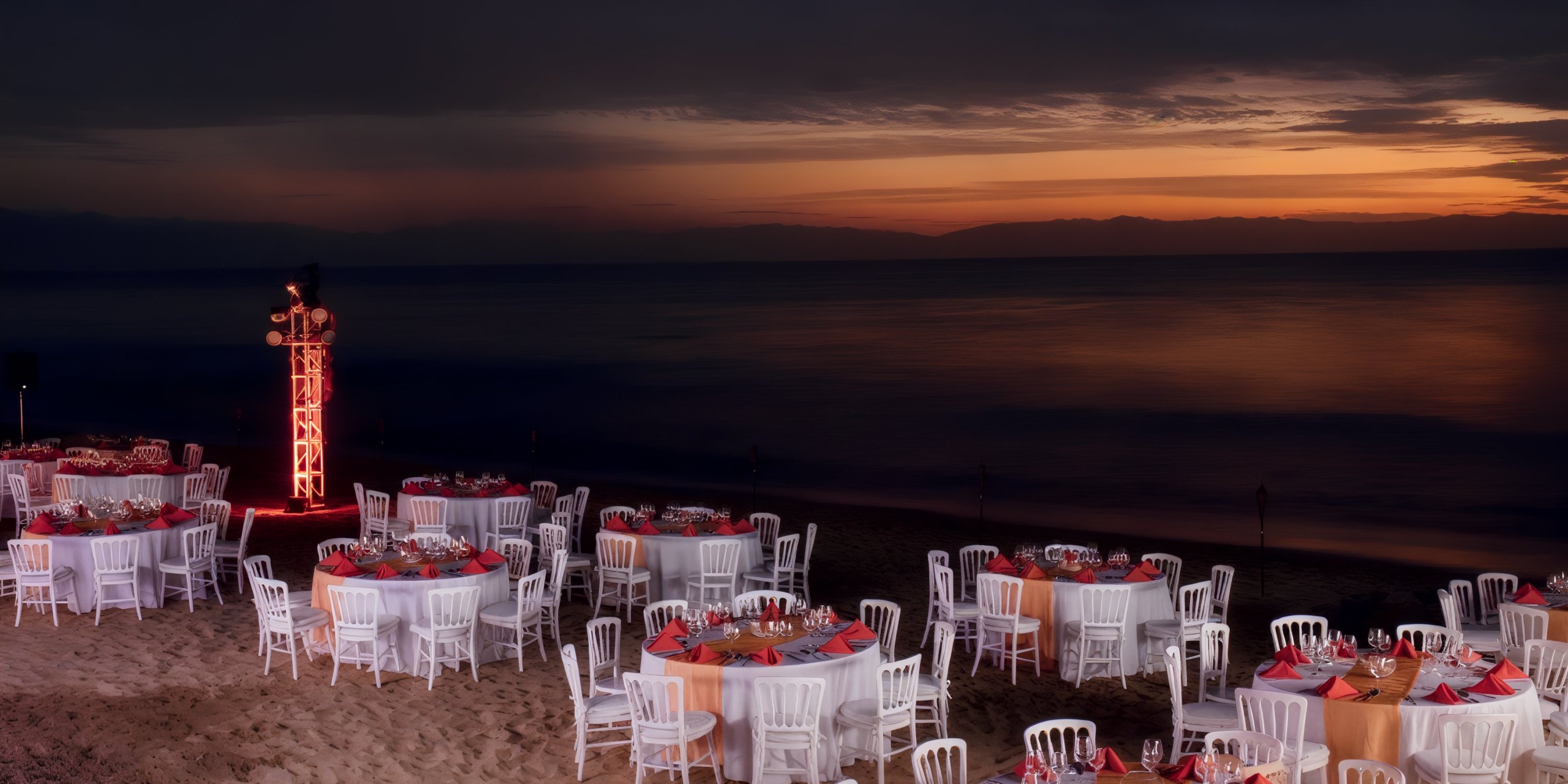Chairs and round tables adorned with red tablecloths are arranged on the beach at sunset, accompanied by an illuminated metal structure nearby at Krystal Grand Nuevo Vallarta.