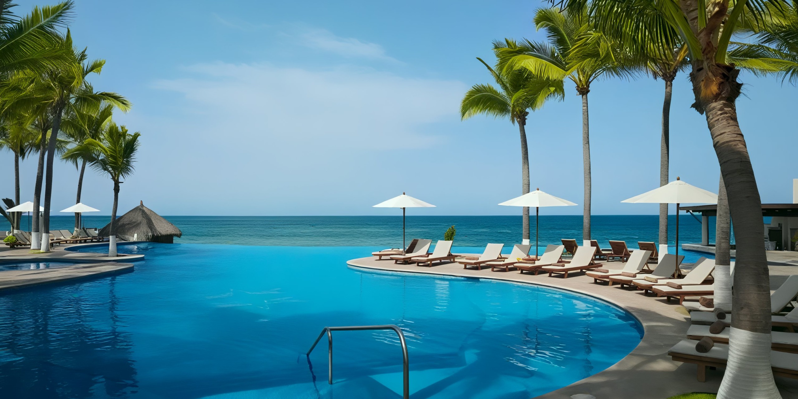 Relax by the infinity pool at Krystal Grand Nuevo Vallarta, where you can enjoy views of the ocean while reclining on lounge chairs with umbrellas nestled under palm trees on a sunny day.