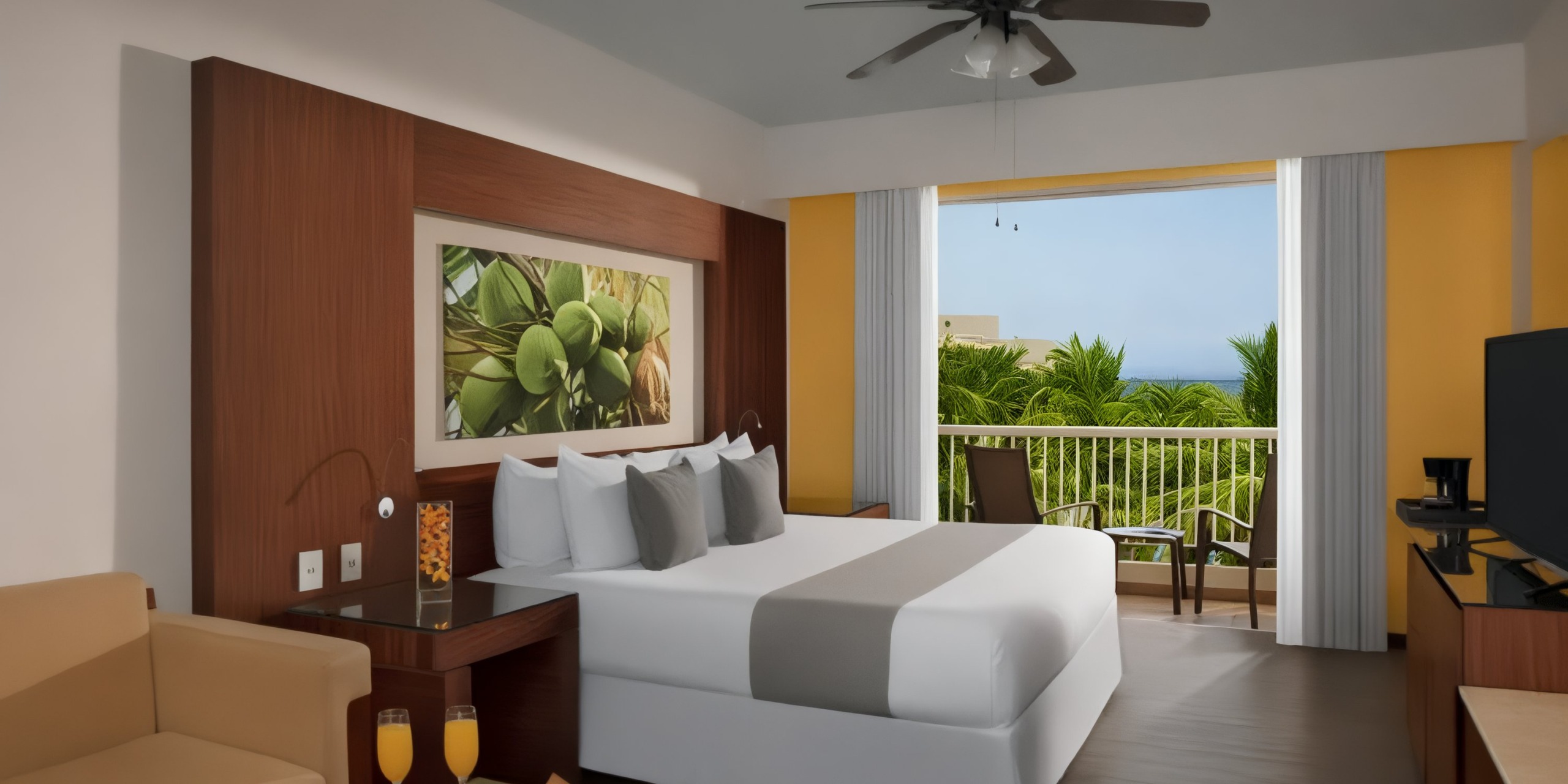 A room at Krystal Grand Nuevo Vallarta offers a king-sized bed, a balcony with a tropical view, and features including a ceiling fan, artwork, a sofa, and a TV. In the foreground sits a table with two glasses of orange juice.