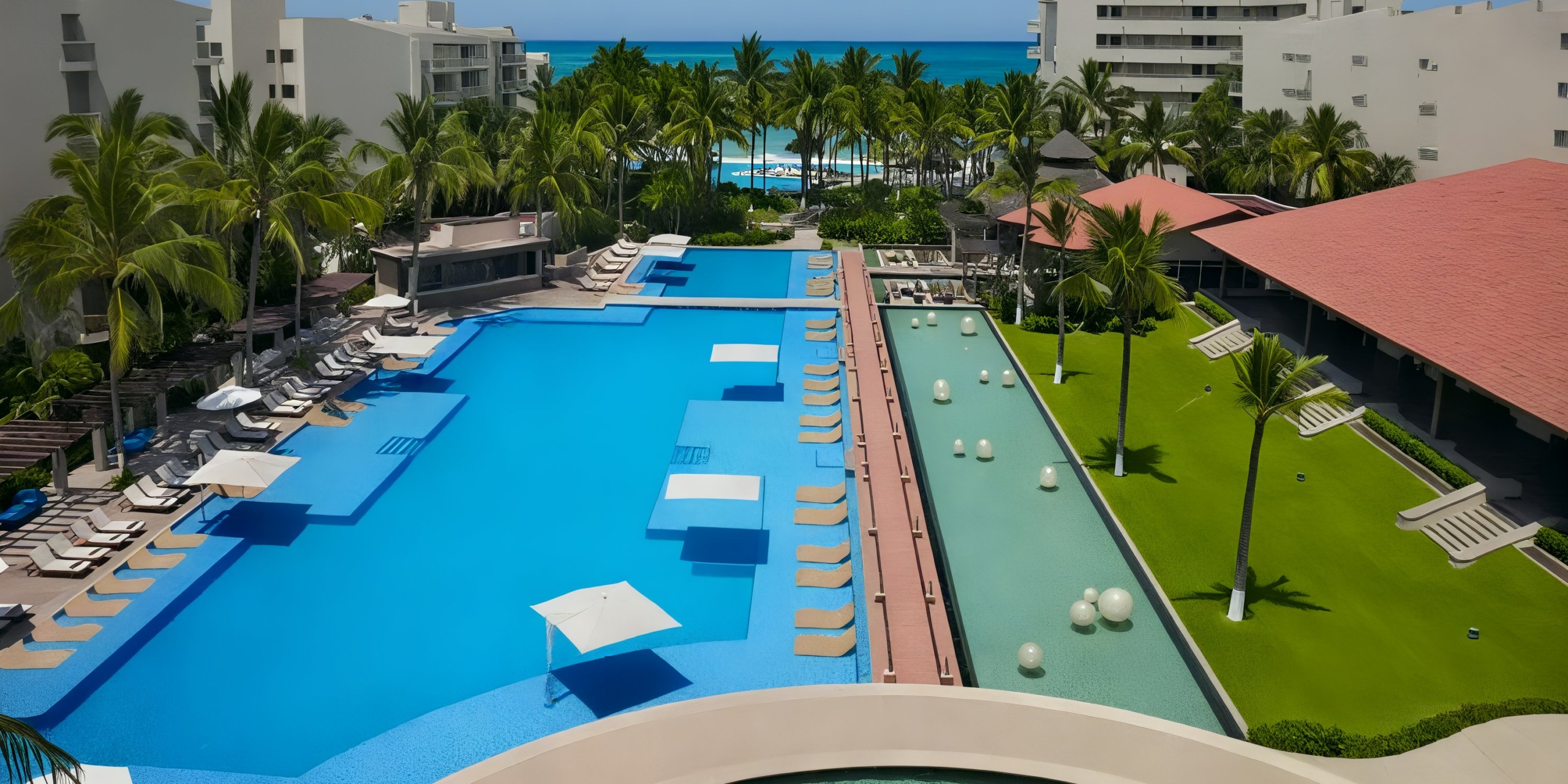 Aerial view of the Krystal Grand Nuevo Vallarta, a luxurious resort with a large pool, sun loungers, palm trees, and a green lawn set against the ocean backdrop.