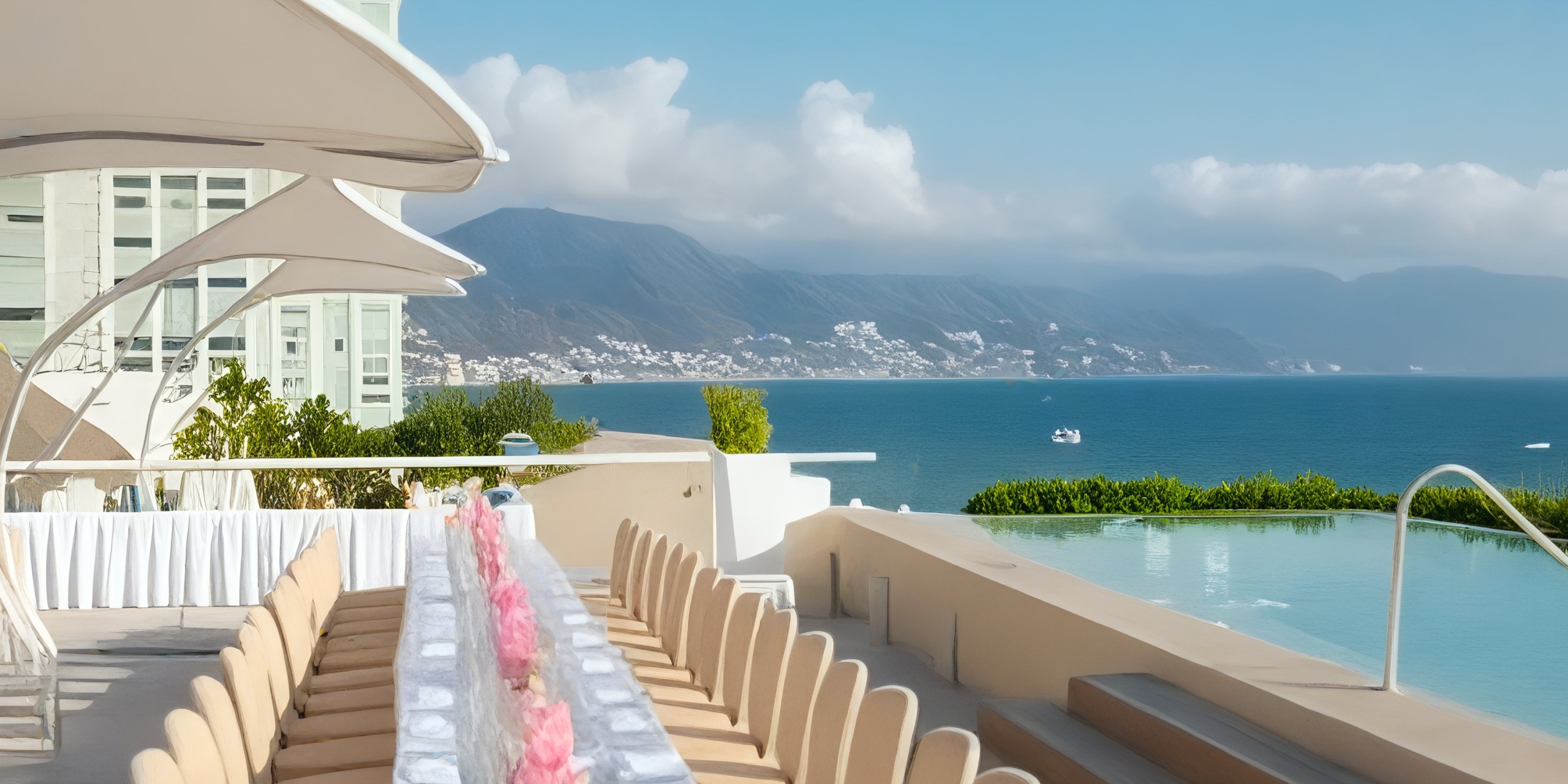 Experience outdoor dining at Krystal Grand Nuevo Vallarta, featuring long tables with pink centerpieces on a terrace that overlooks an infinity pool and offers a breathtaking view of the ocean, majestic mountains, and a cloudy sky.