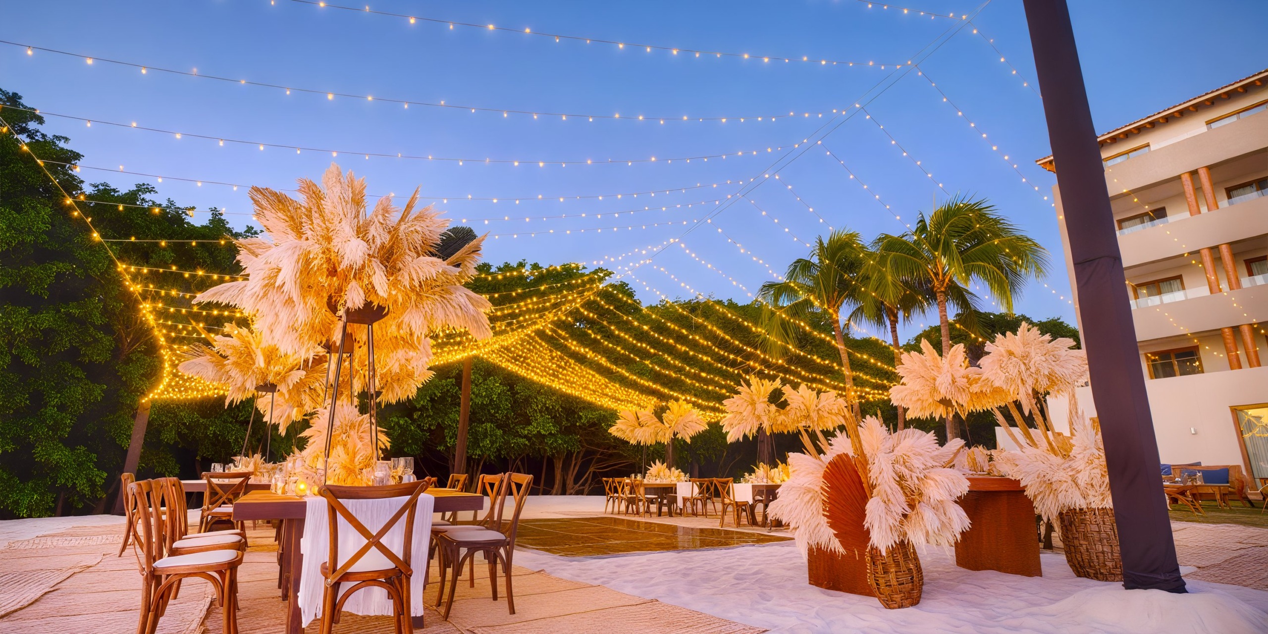 The outdoor dining area at Marival Armony, set on a sandy beach under a clear evening sky, is adorned with string lights and pampas grass arrangements.