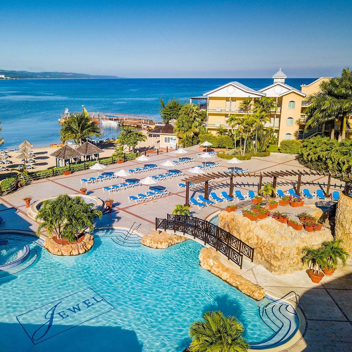 Aerial view of Jewel Paradise Cove with a large swimming pool, sun loungers, and a pier extending into a calm blue sea under a clear sky.