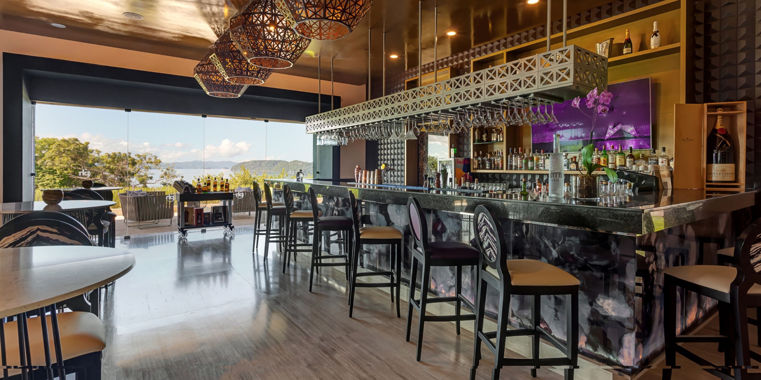 Step into the modern interior of Planet Hollywood Costa Rica, featuring a sleek counter, comfortable high chairs, stylish pendant lights, and a large window offering a breathtaking scenic view. Bottles and glasses are neatly arranged on shelves and the counter for a polished look.