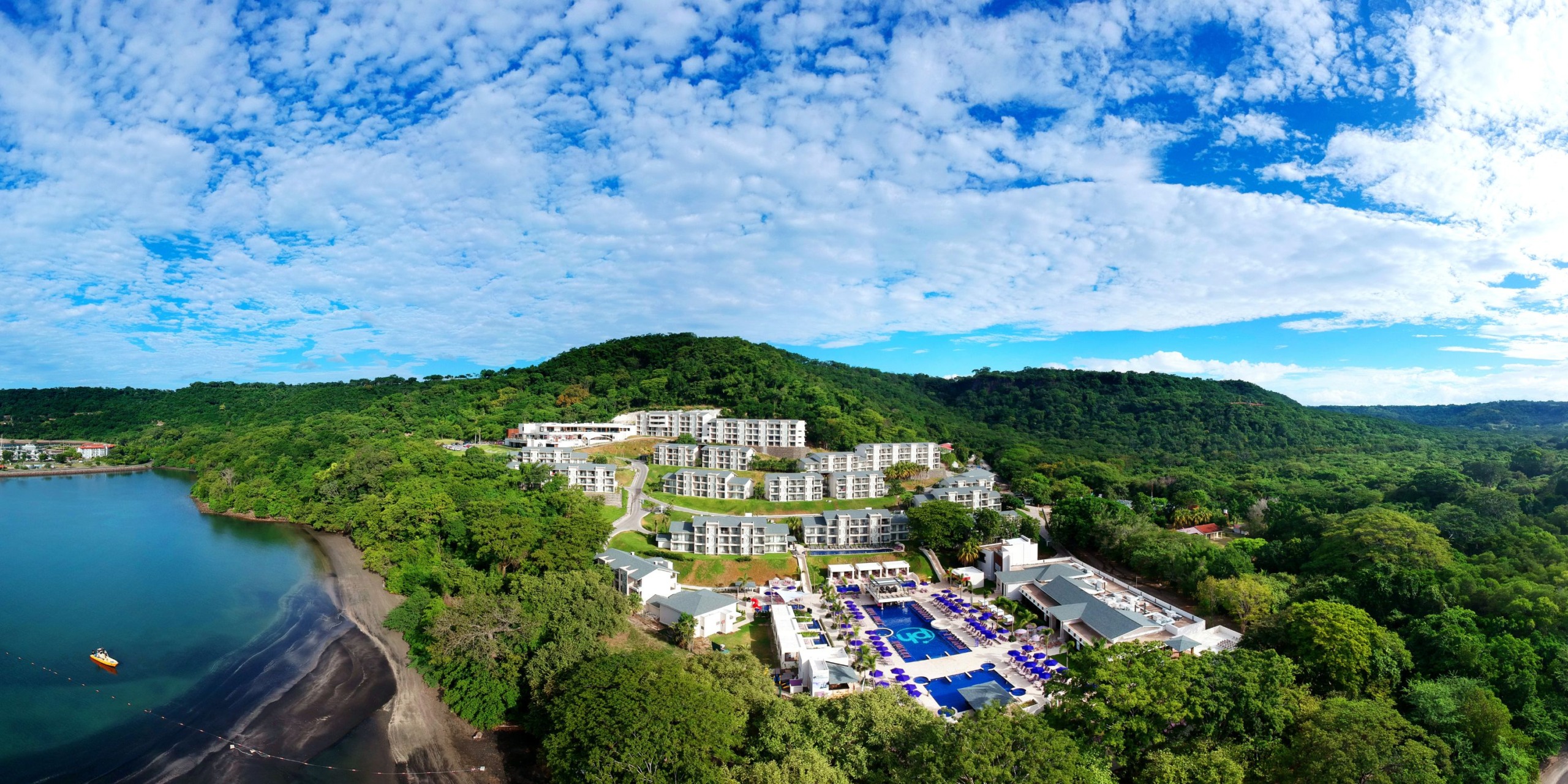 Bird's-eye view of Planet Hollywood Costa Rica, a coastal resort enveloped by vibrant greenery and a sandy beach, featuring multiple buildings and pools beneath a partly cloudy blue sky.
