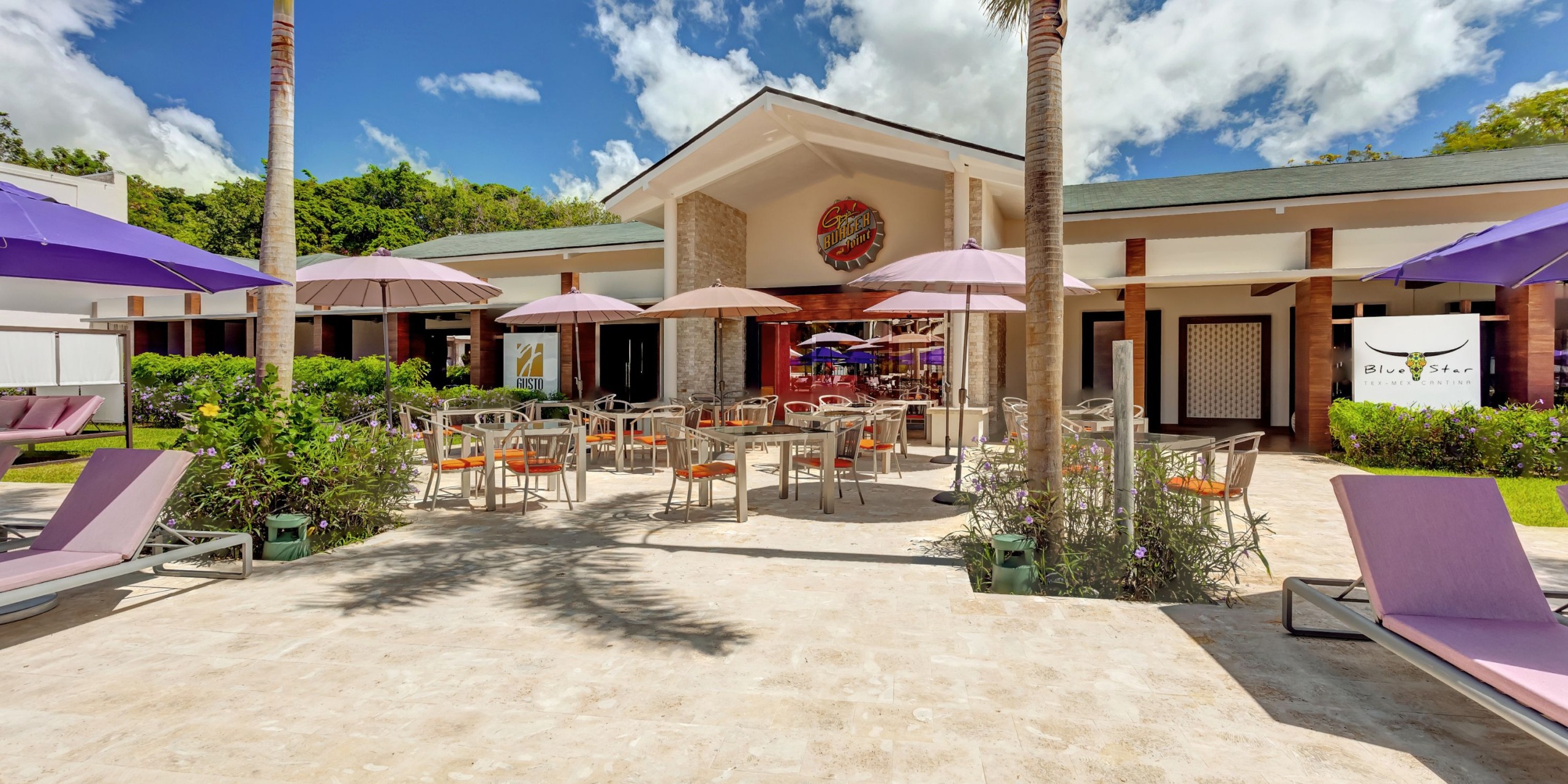 Planet Hollywood Costa Rica features an outdoor dining area with tables, chairs, and purple umbrellas on a sunny day, located in front of the establishment's sign and enveloped by lush greenery.