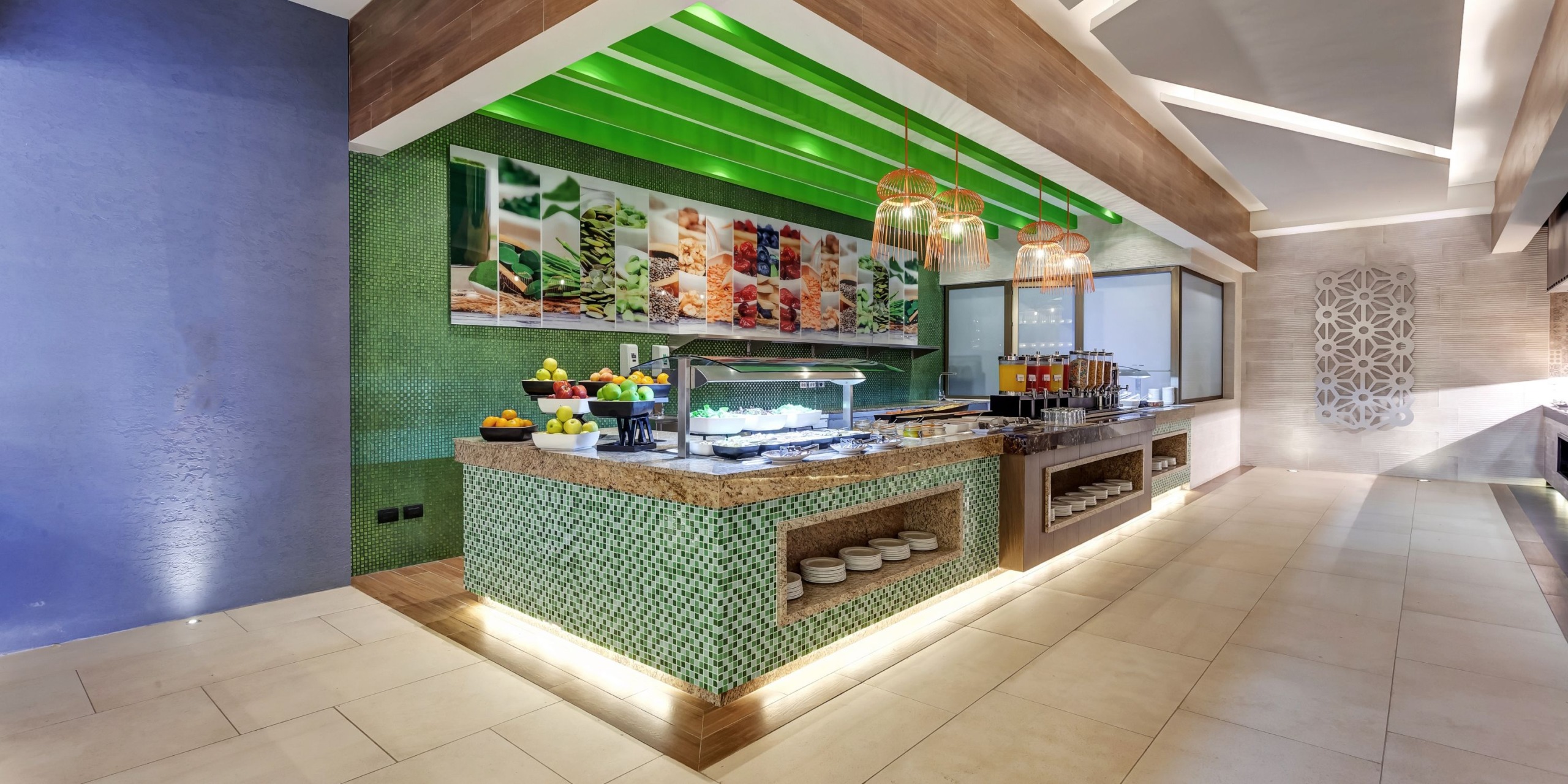 A contemporary buffet arrangement featuring a range of vibrant dishes and drinks is showcased on a green-tiled counter beneath a green accent ceiling at Planet Hollywood Costa Rica.