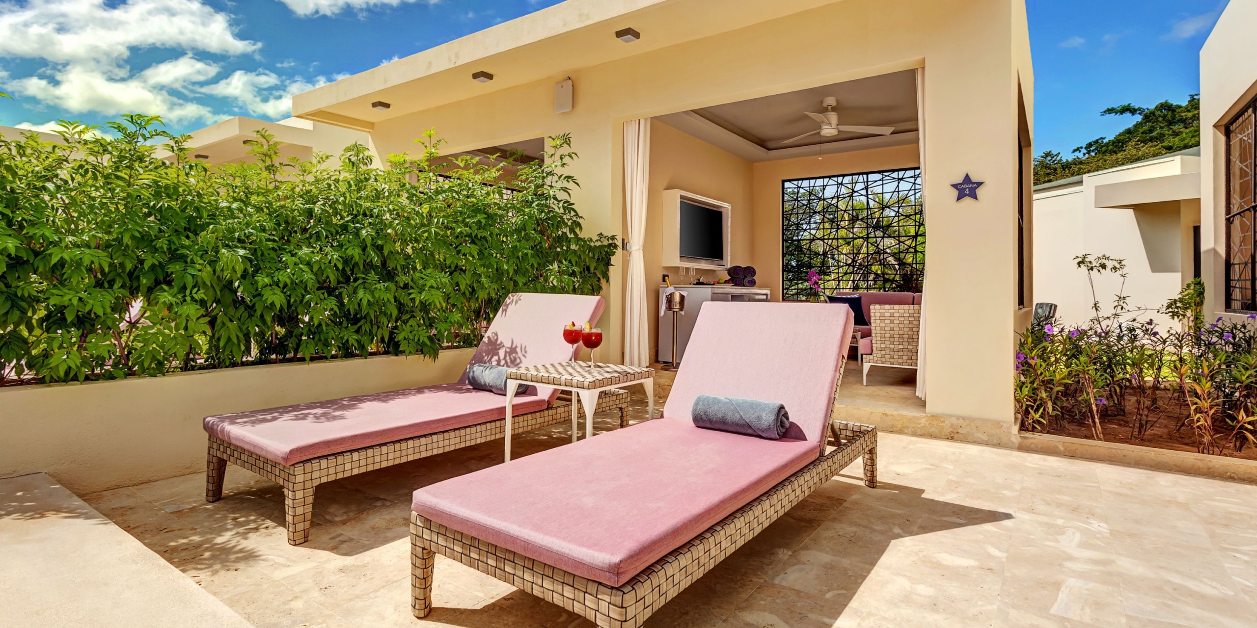 Outdoor patio at Planet Hollywood Costa Rica featuring two pink loungers facing a small table, bordered by a green hedge. A room in the background is equipped with a TV and ceiling fan, with clear skies above.