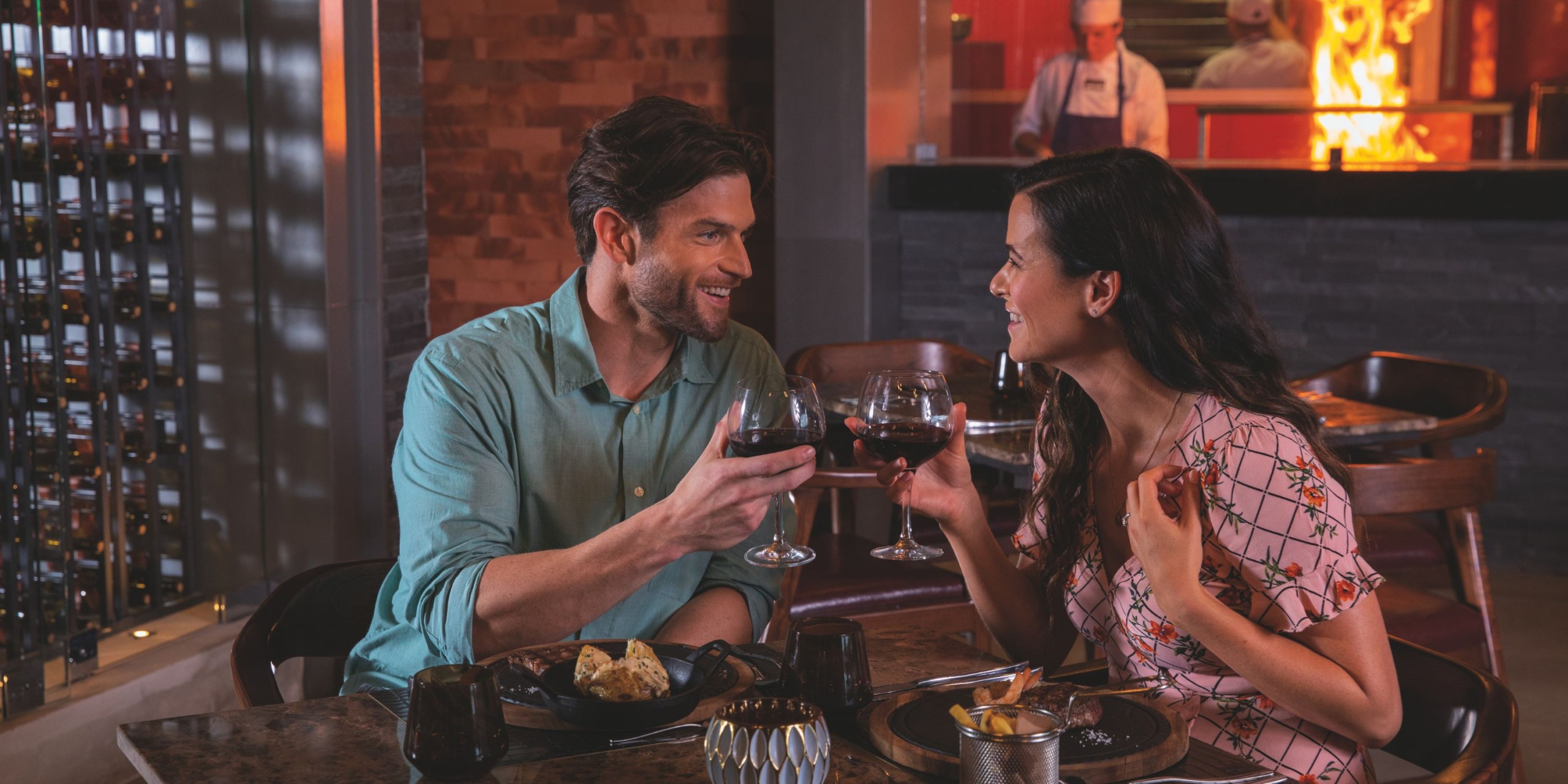 A couple clinks wine glasses while dining at Planet Hollywood Costa Rica, with a chef cooking in the background.