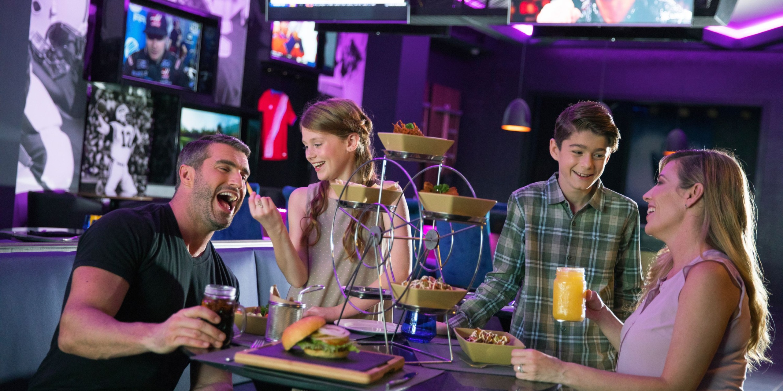 Four people enjoy food and drinks at a table with a Ferris wheel-shaped snack holder in Planet Hollywood Costa Rica, a sports-themed restaurant.