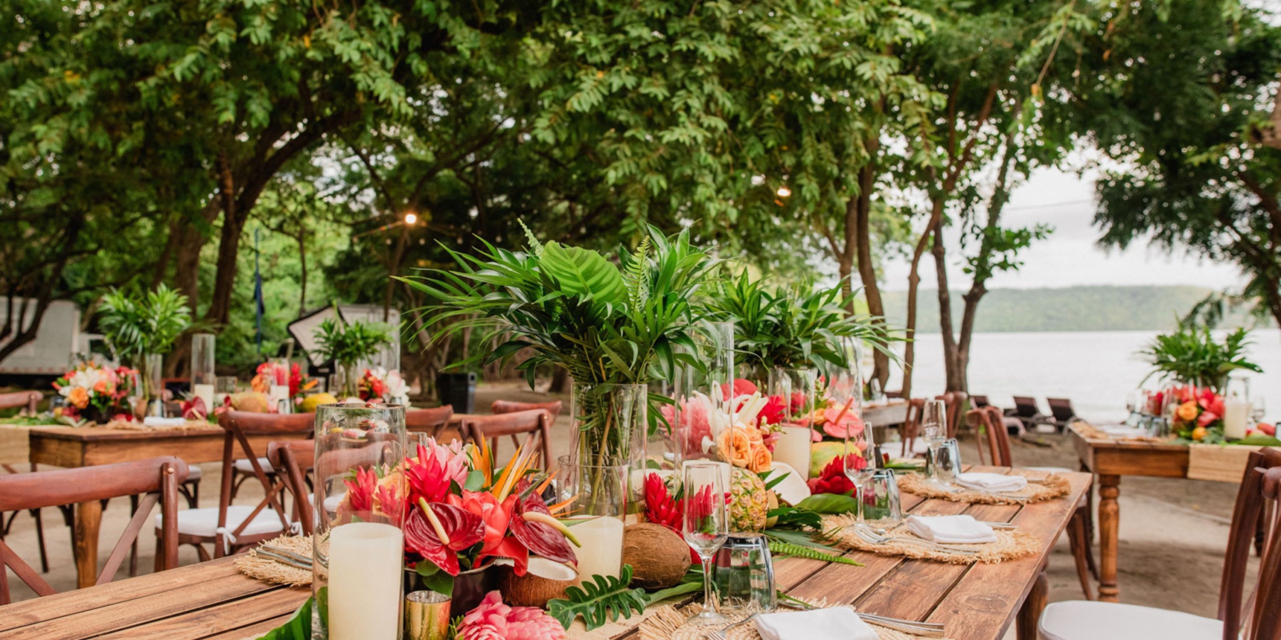 Experience outdoor dining at Planet Hollywood Costa Rica, featuring wooden tables adorned with tropical floral centerpieces and surrounded by lush greenery in a forested area near a tranquil body of water.