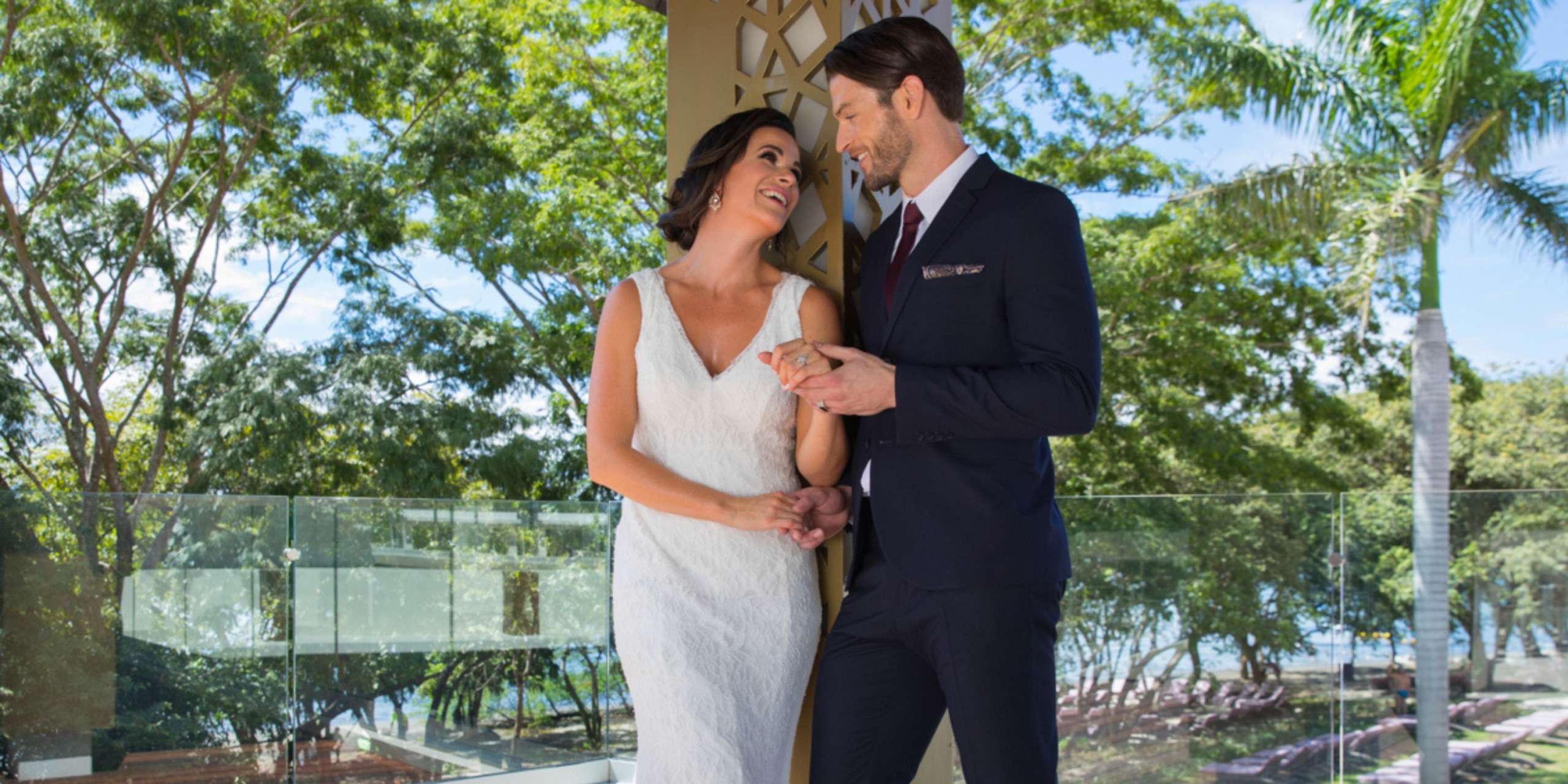 A bride in a white dress and a groom in a dark suit stand together, holding hands and smiling against the lush, tree-filled backdrop of Planet Hollywood Costa Rica.