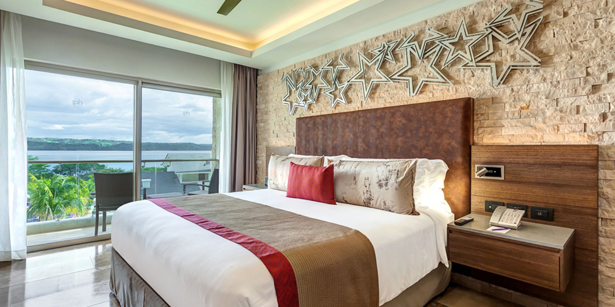 Experience the Planet Hollywood Costa Rica, featuring a contemporary room equipped with a spacious bed, stylish red and brown accents, star-shaped wall decor, and a balcony offering scenic views of lush trees and shimmering water.