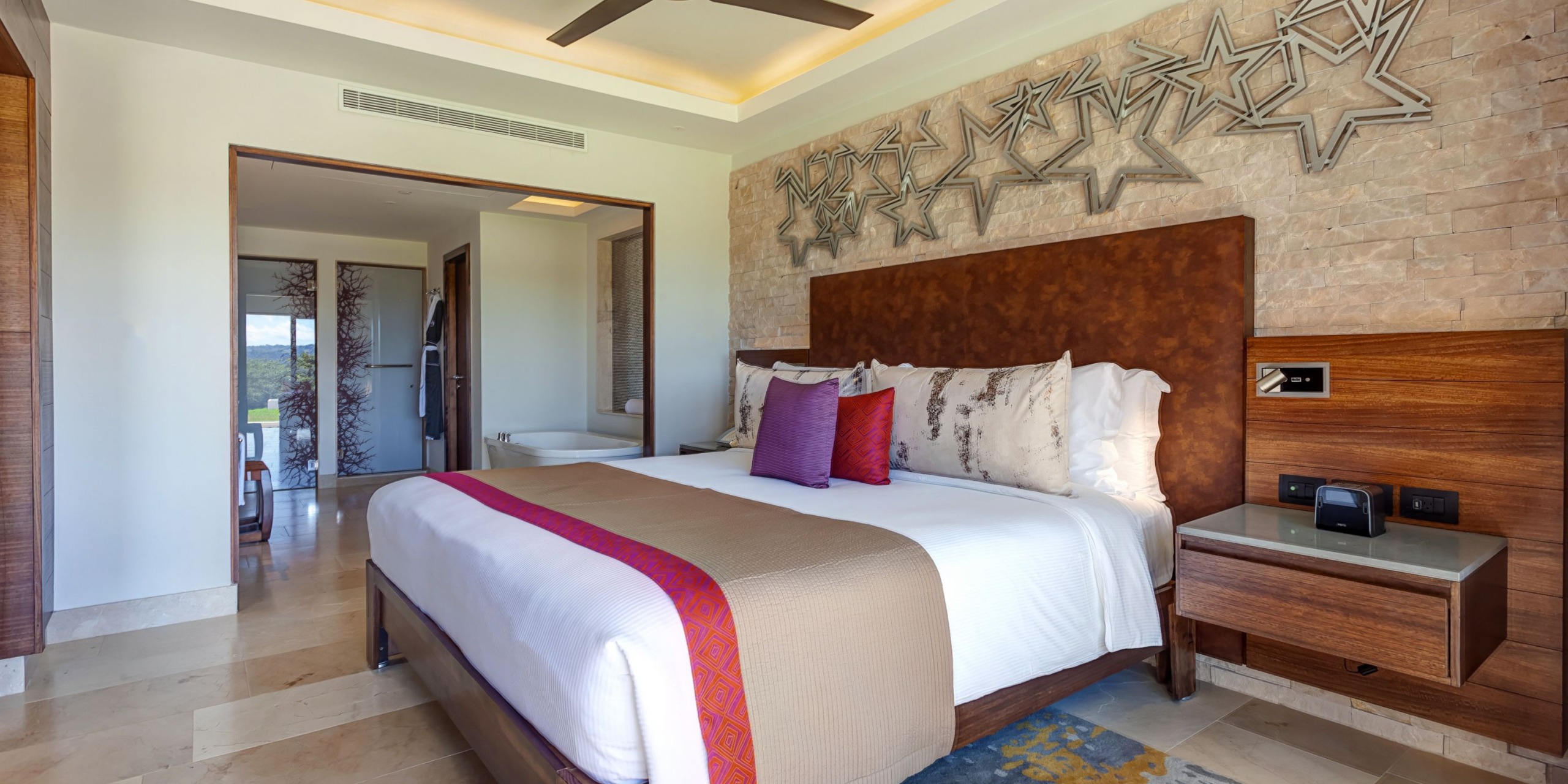 Modern bedroom at Planet Hollywood Costa Rica featuring a king-sized bed, star-shaped wall decorations, wooden furniture, and a view into the bathroom.