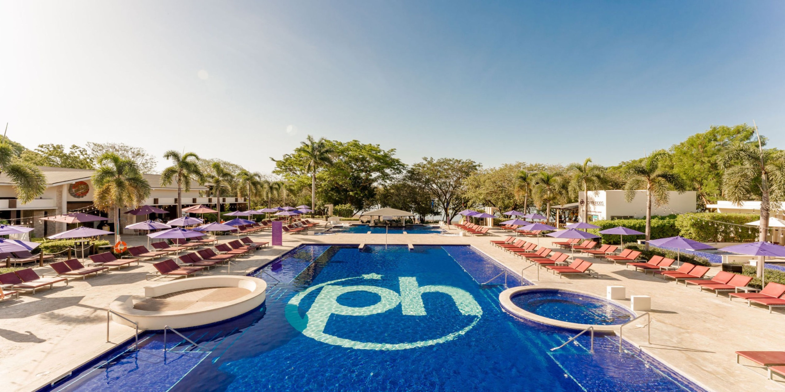 The resort pool area at Planet Hollywood Costa Rica features the signature "ph" logo, surrounded by palm trees, lounge chairs, and vibrant purple umbrellas set against a clear blue sky.