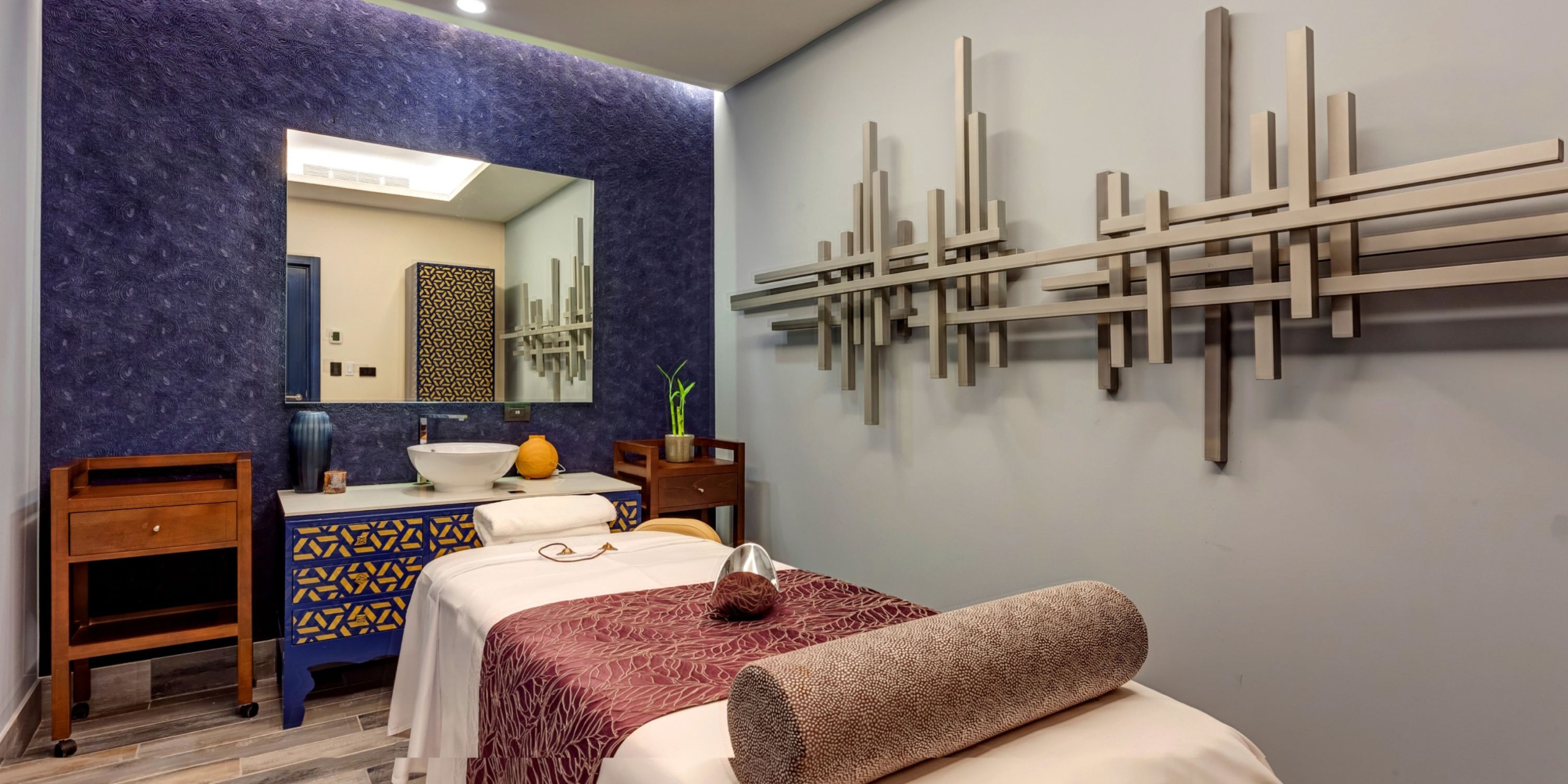Experience the Planet Hollywood Costa Rica with its tranquil massage room, featuring a treatment table, pillow, and towel under beautiful decorative wall art. In the background, you'll find a sink paired with a mirror and a charming table adorned with a small vase containing green leaves.