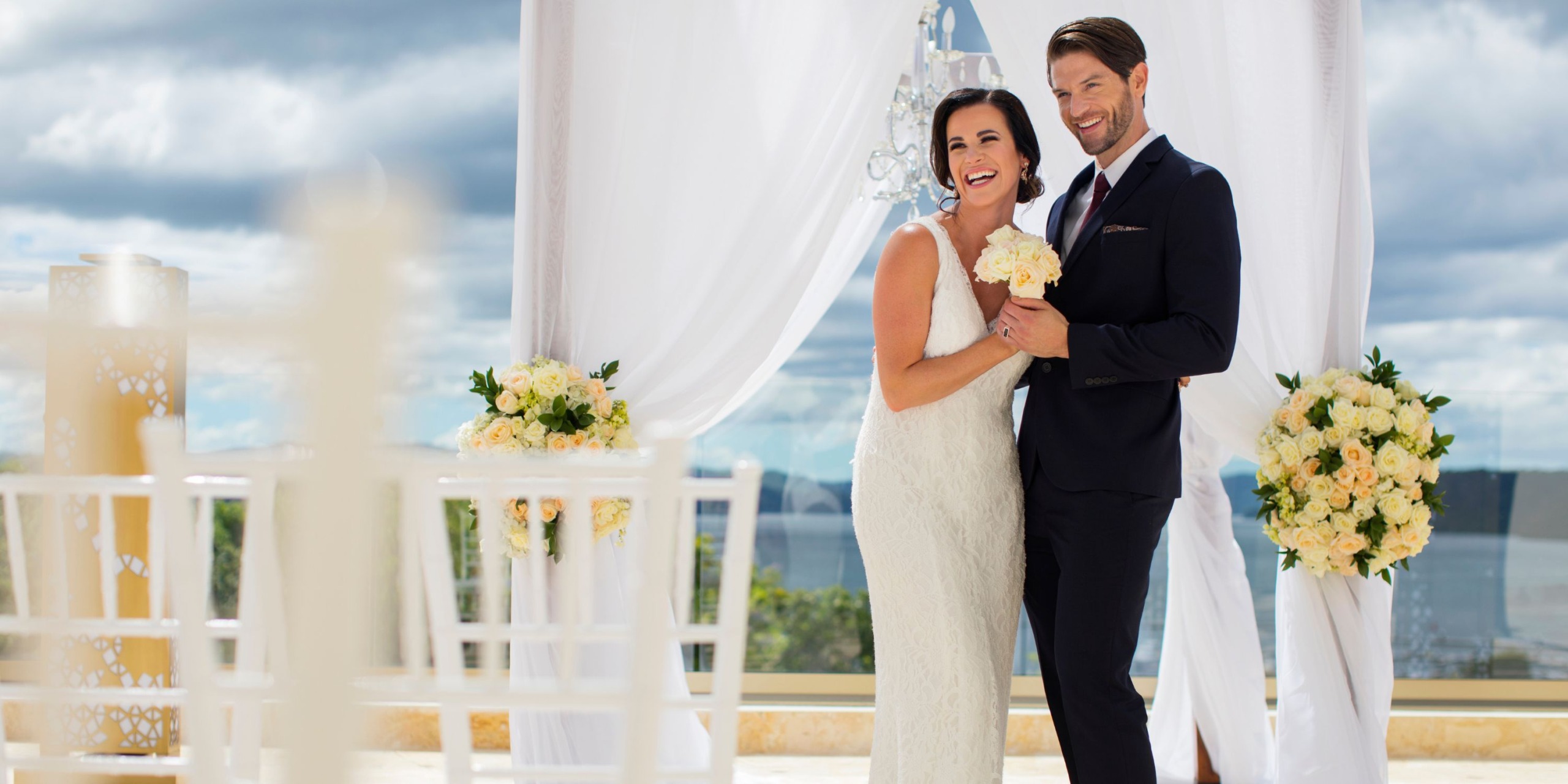 A couple in wedding attire smiles in front of a white draped arch adorned with flowers at Planet Hollywood Costa Rica.