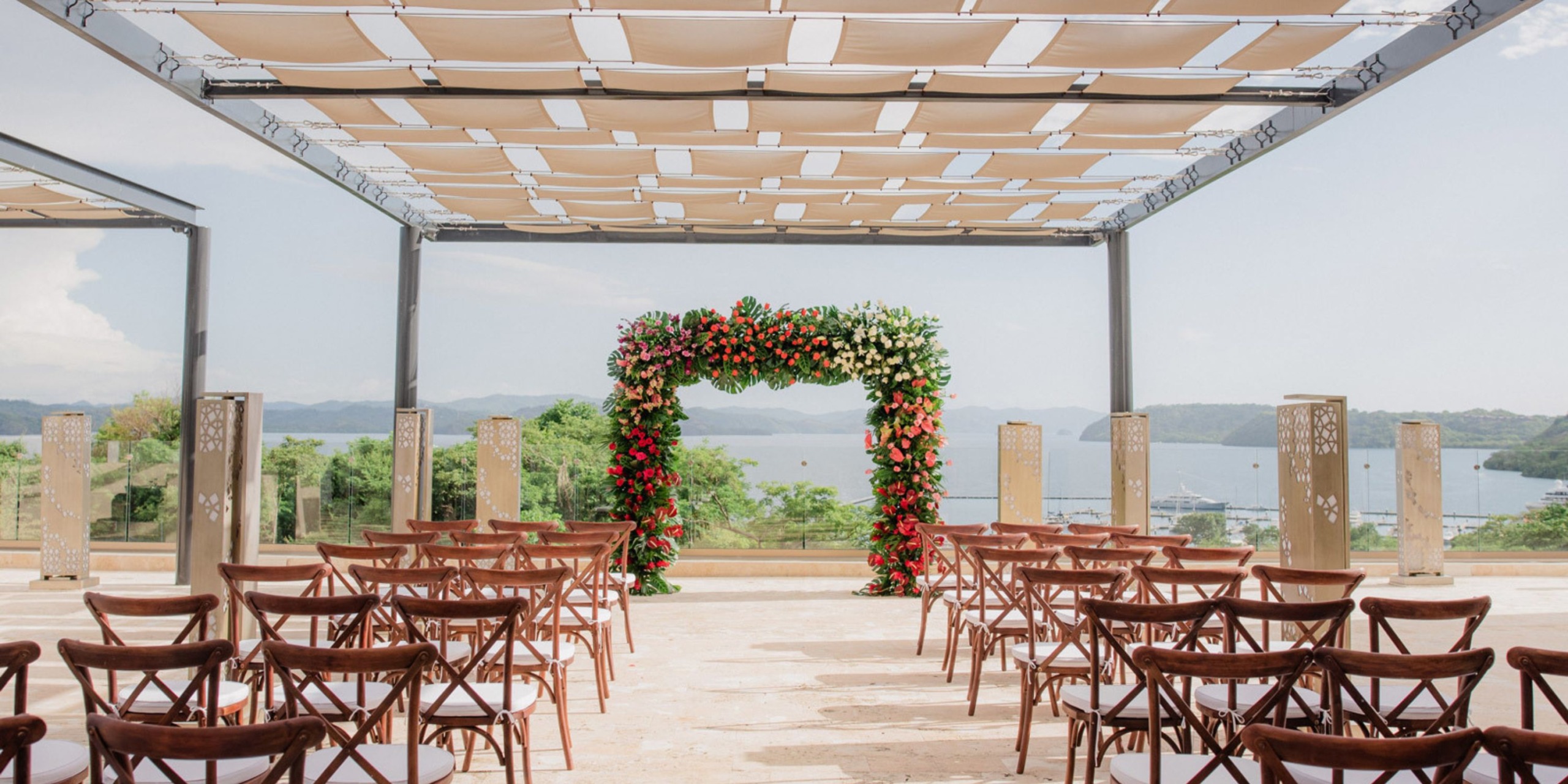 At Planet Hollywood Costa Rica, you can have an outdoor wedding setup featuring wooden chairs directed towards a floral arch beneath a canopy, with breathtaking views of the scenic landscape, water, and hills in the background.