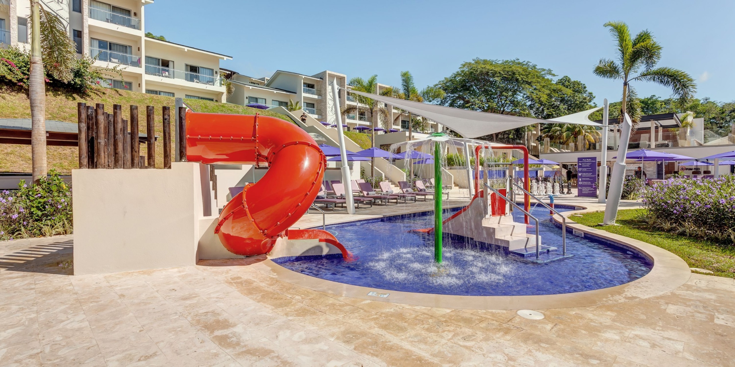 Planet Hollywood Costa Rica features an outdoor water play area that includes a vibrant red slide, a small splash pool, and shaded seating. The area is beautifully surrounded by lush greenery and nearby buildings.