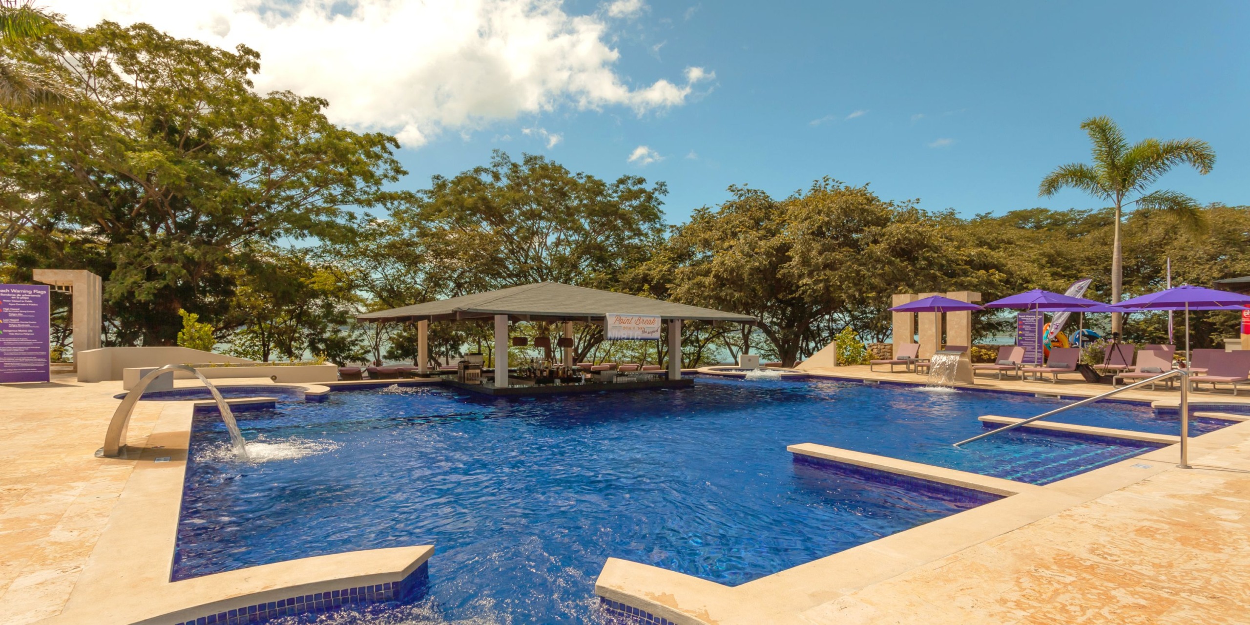 Enjoy the outdoor swimming pool at Planet Hollywood Costa Rica, featuring a bar at its center and surrounded by lush trees and lounge chairs shaded by purple umbrellas.