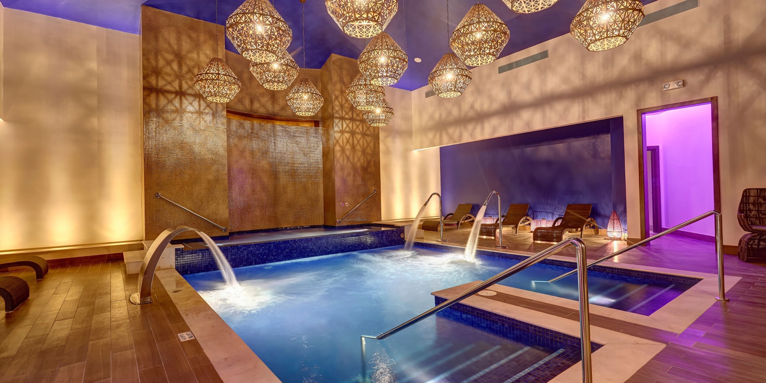 Luxurious indoor pool area at Planet Hollywood Costa Rica, featuring cascading water features and ornate hanging lights. Comfortable lounge chairs rest on a wooden deck, while a lit, purple doorway can be seen to the right.
