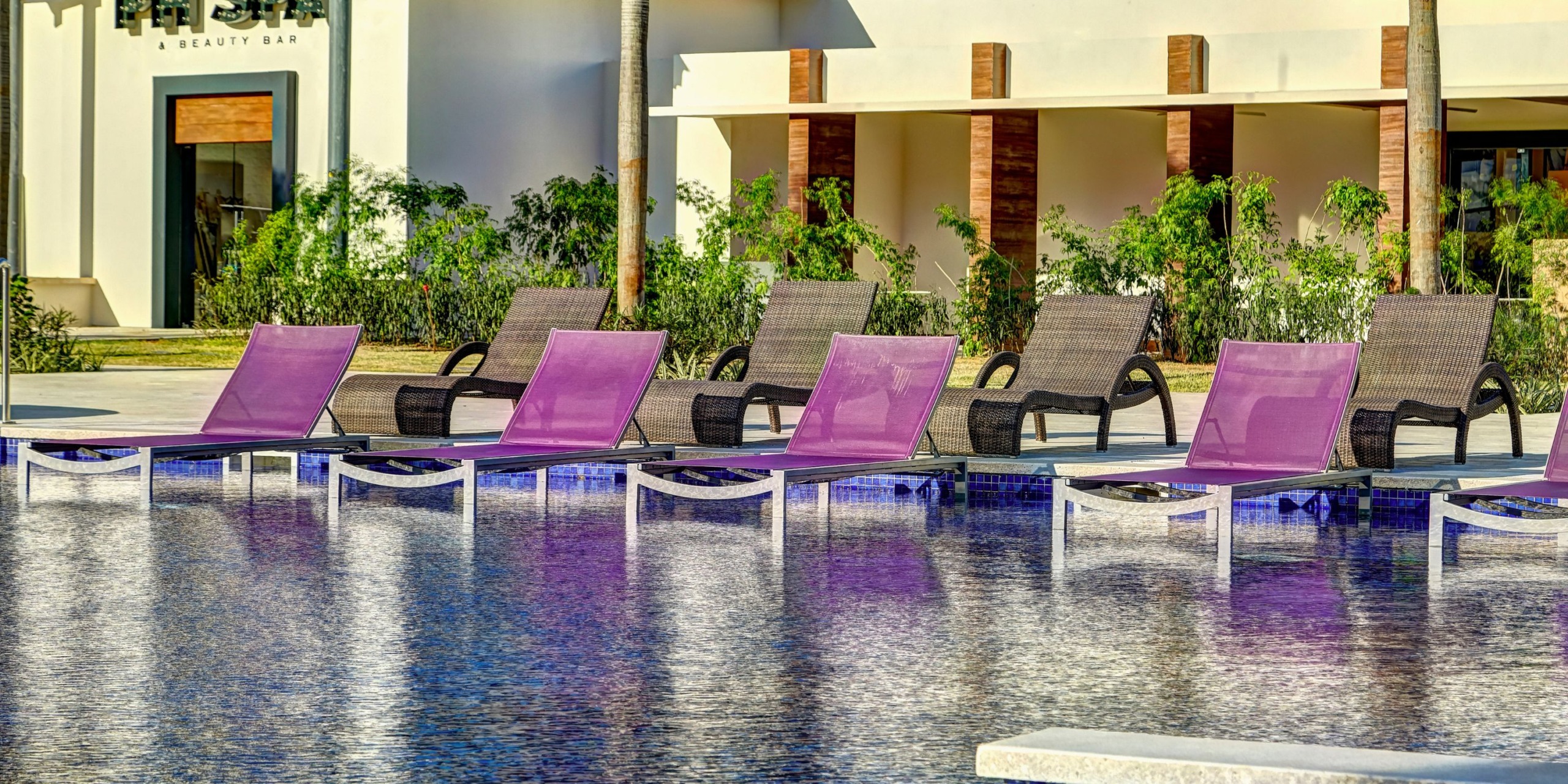 Planet Hollywood Costa Rica features purple and brown lounge chairs by a poolside, framed by a building and lush greenery in the background.