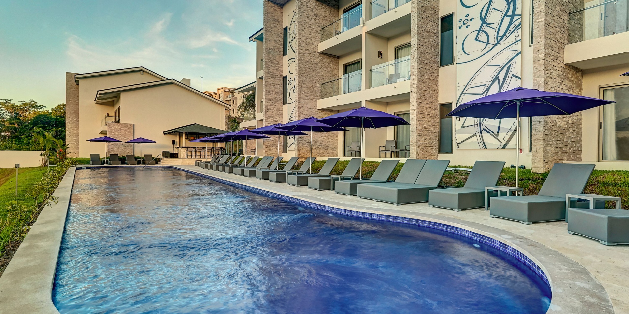 The Planet Hollywood Costa Rica features an outdoor pool adorned with lounge chairs and vibrant purple umbrellas, set against a modern architectural backdrop.