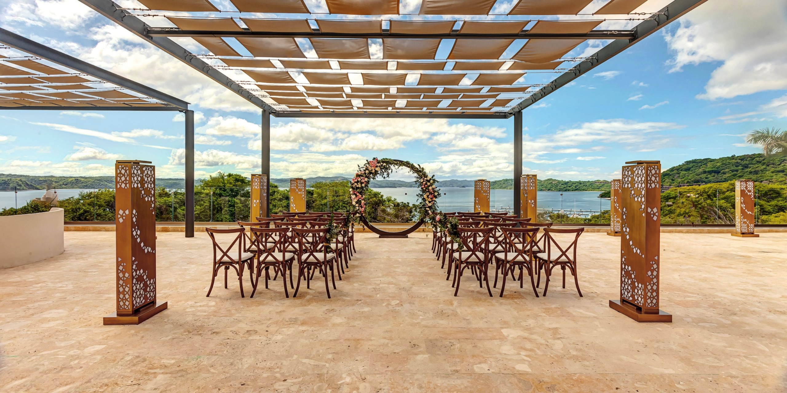 Outdoor wedding setup at Planet Hollywood Costa Rica with rows of wooden chairs facing a circular floral arch. The venue offers a scenic view of trees and water under a partly cloudy sky.