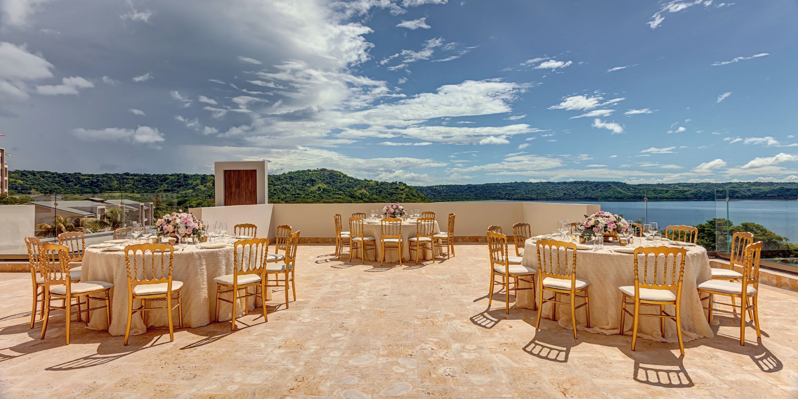 The outdoor terrace at Planet Hollywood Costa Rica is elegantly arranged for an event, showcasing round tables adorned with floral centerpieces and surrounded by wooden chairs. Guests can enjoy a scenic view of the tranquil lake and lush green hills under a partly cloudy sky.