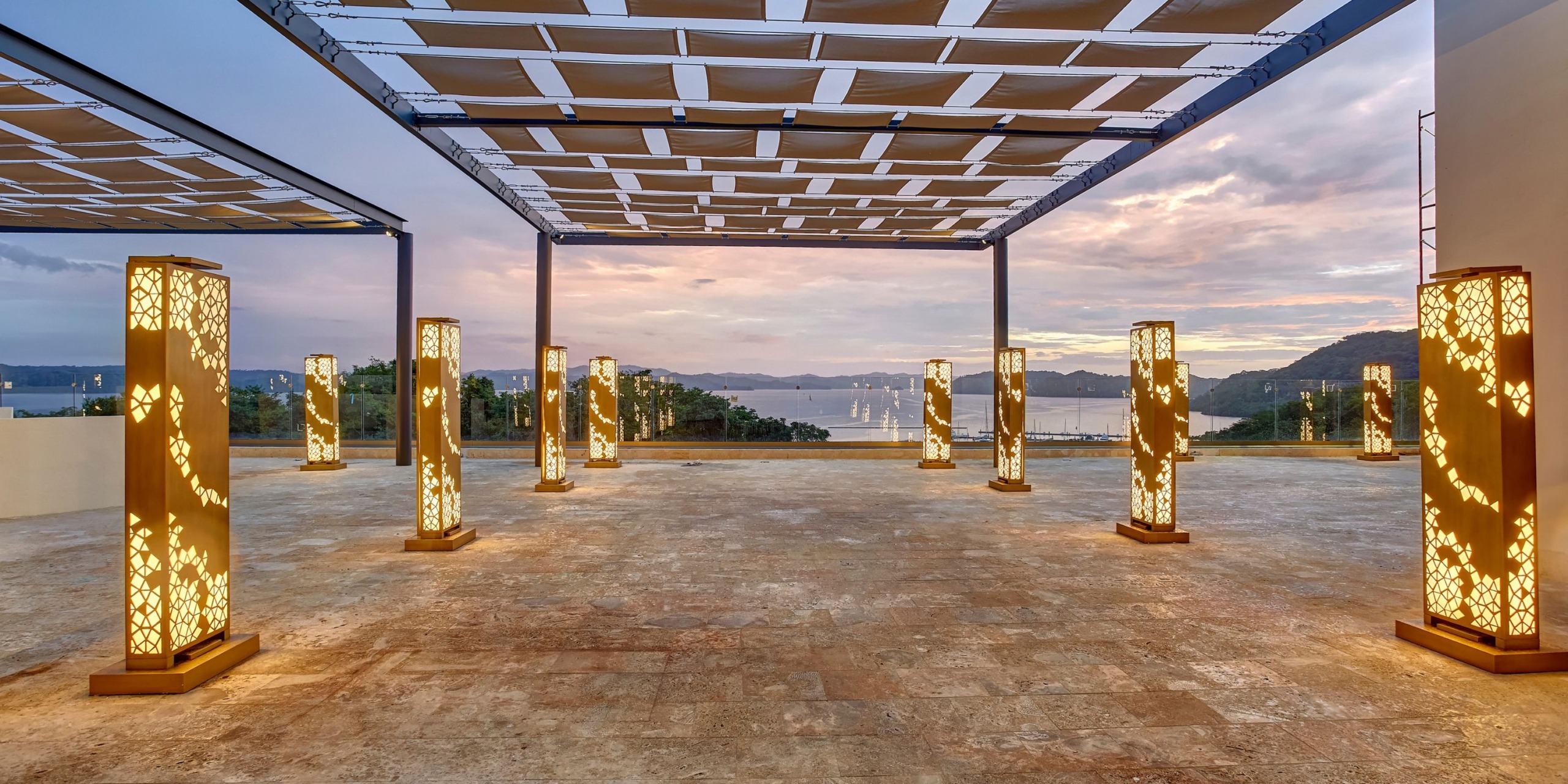 Outdoor terrace at Planet Hollywood Costa Rica with illuminated decorative pillars and a slatted roof, offering a scenic sunset view of distant hills and water.