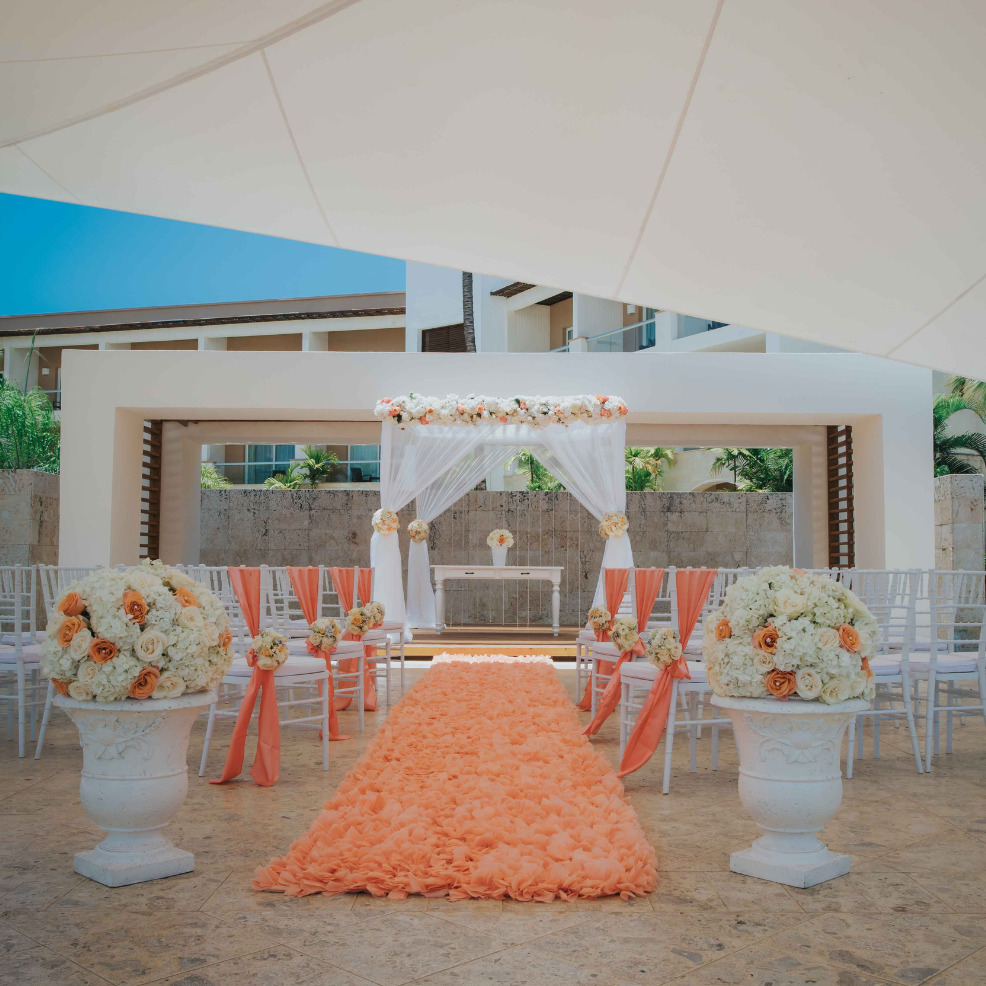 Destination wedding setup with a floral arch, orange aisle runner, and white chairs under large umbrellas.