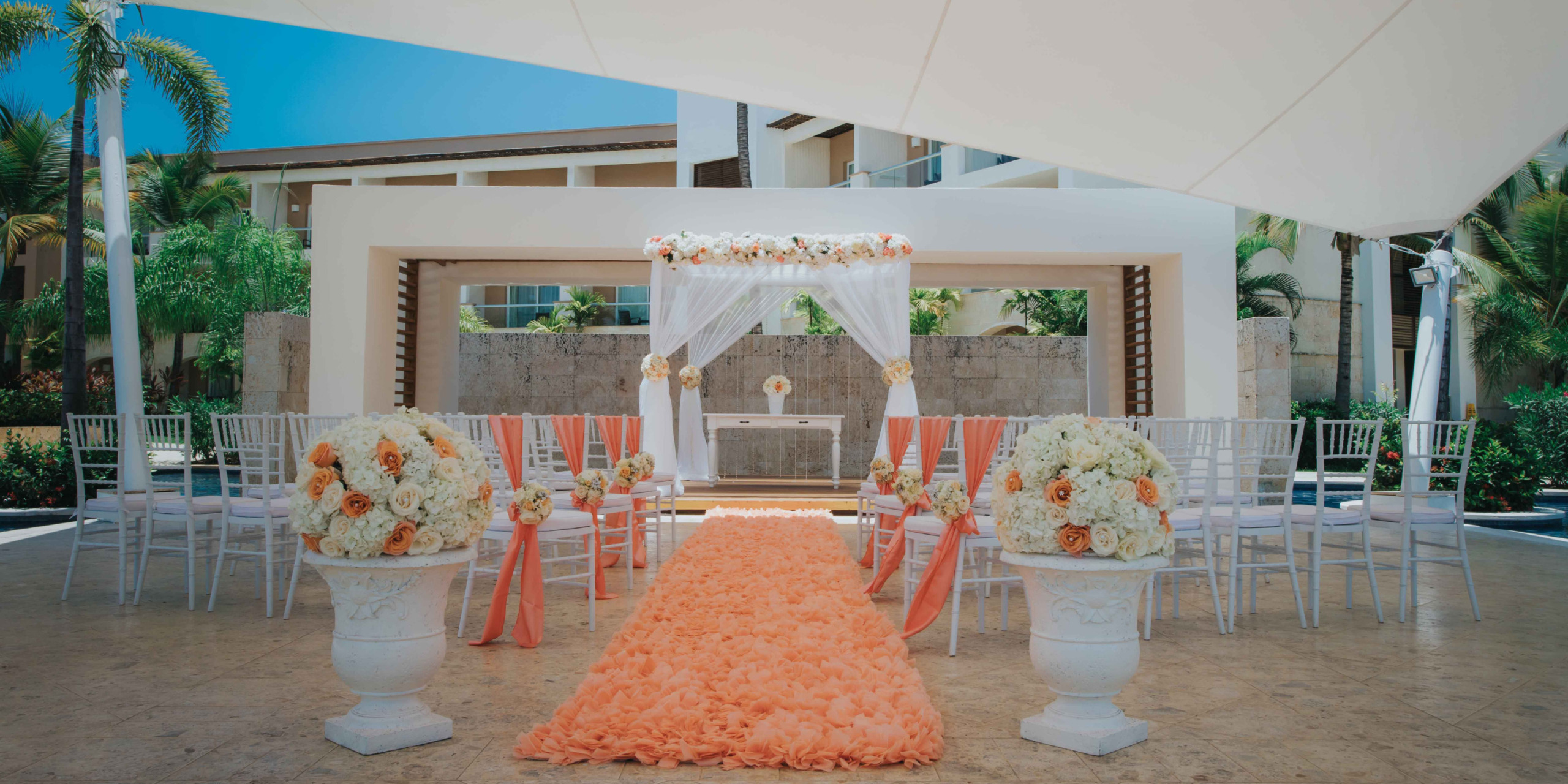 Destination wedding setup with a floral decorated aisle, white chairs, and a canopy under large umbrellas, surrounded by palm trees.