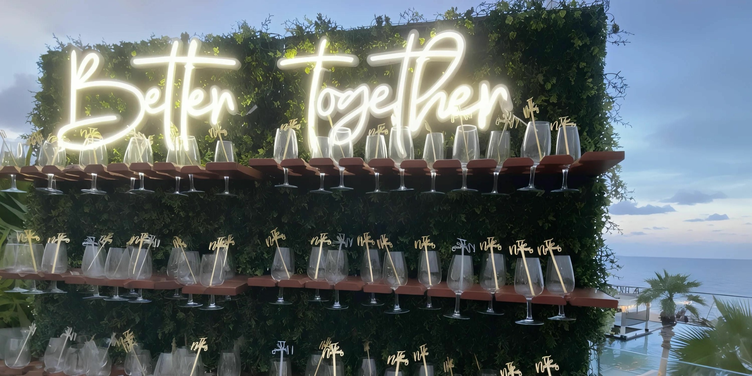 A greenery-covered wall, adorned with neon letters spelling "Better Together," holds numerous Champagne glasses each labeled with a name, overlooking the scenic ocean view at Secrets Riviera Cancun.