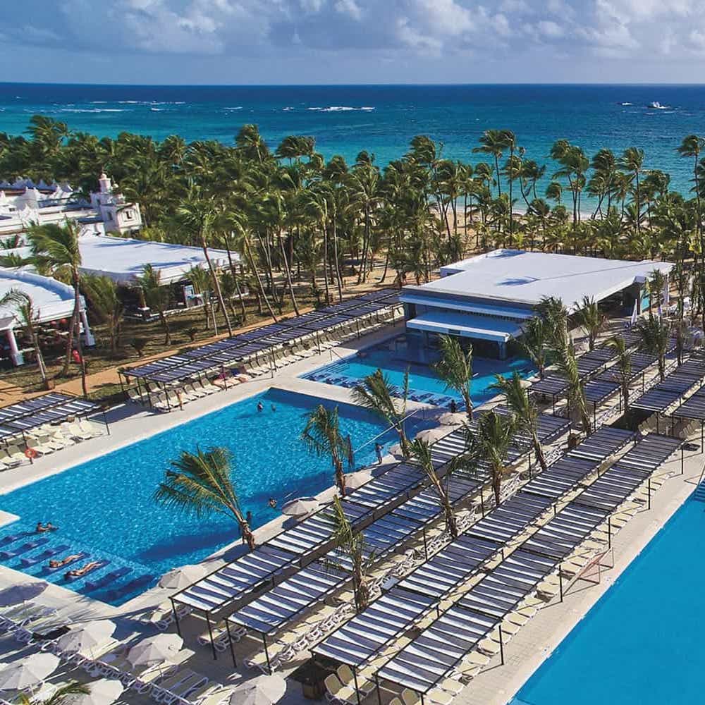 Aerial view of Hotel Riu Bambu, a tropical resort destination for weddings with a large swimming pool, palm trees, white beachfront villas, and a vivid blue sea in the background.