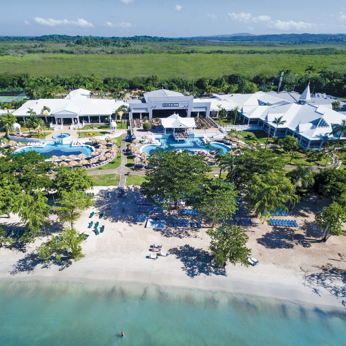 Aerial view of Hotel Riu Negril, a tropical resort specializing in destination weddings, with white buildings and swimming pools near a sandy beach, surrounded by lush greenery and clear blue waters.